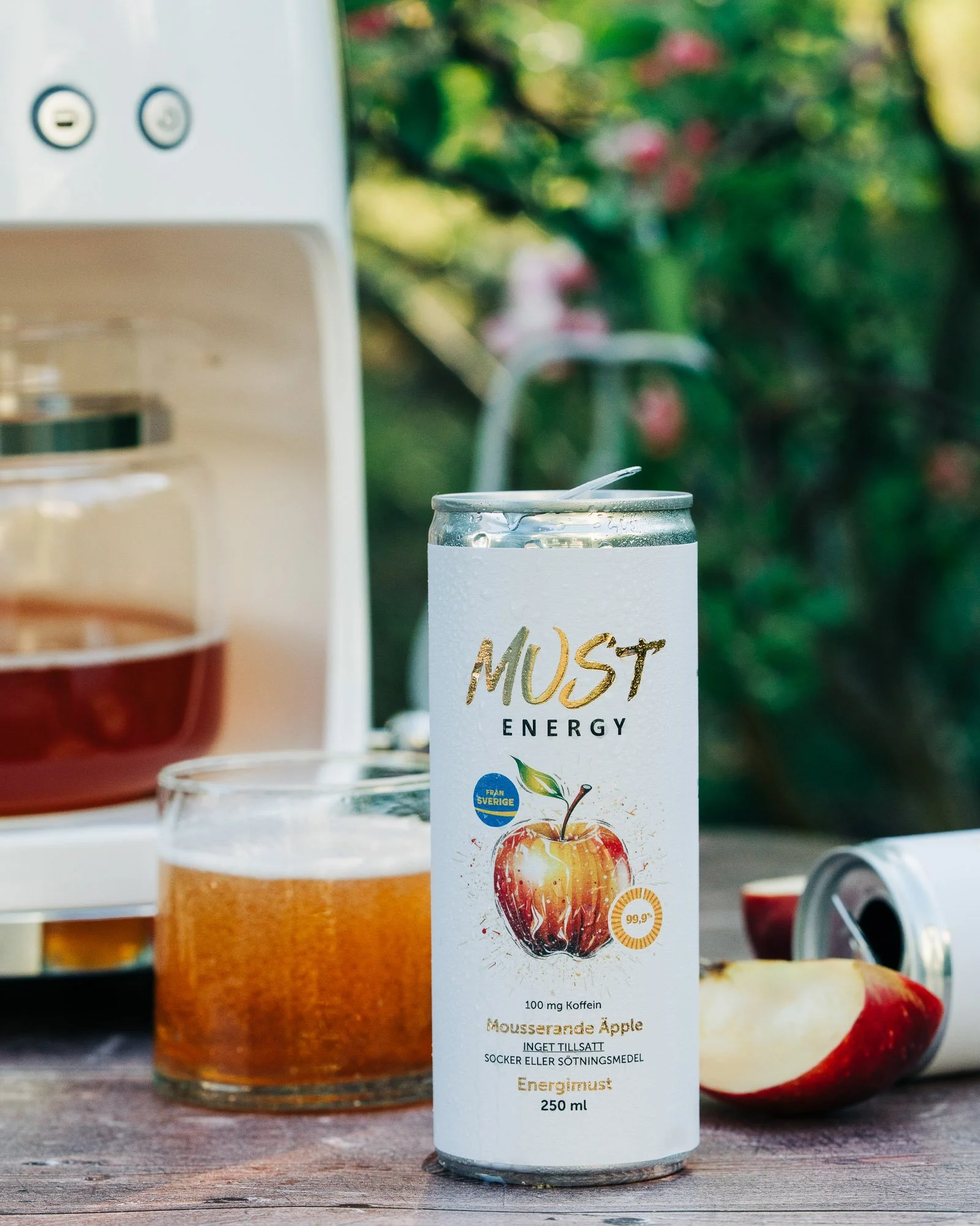 A can of energy drink labeled 'Must Energy' with an apple graphic and information about caffeine content, on a wooden surface outdoors with blurred background of greenery and flowers.