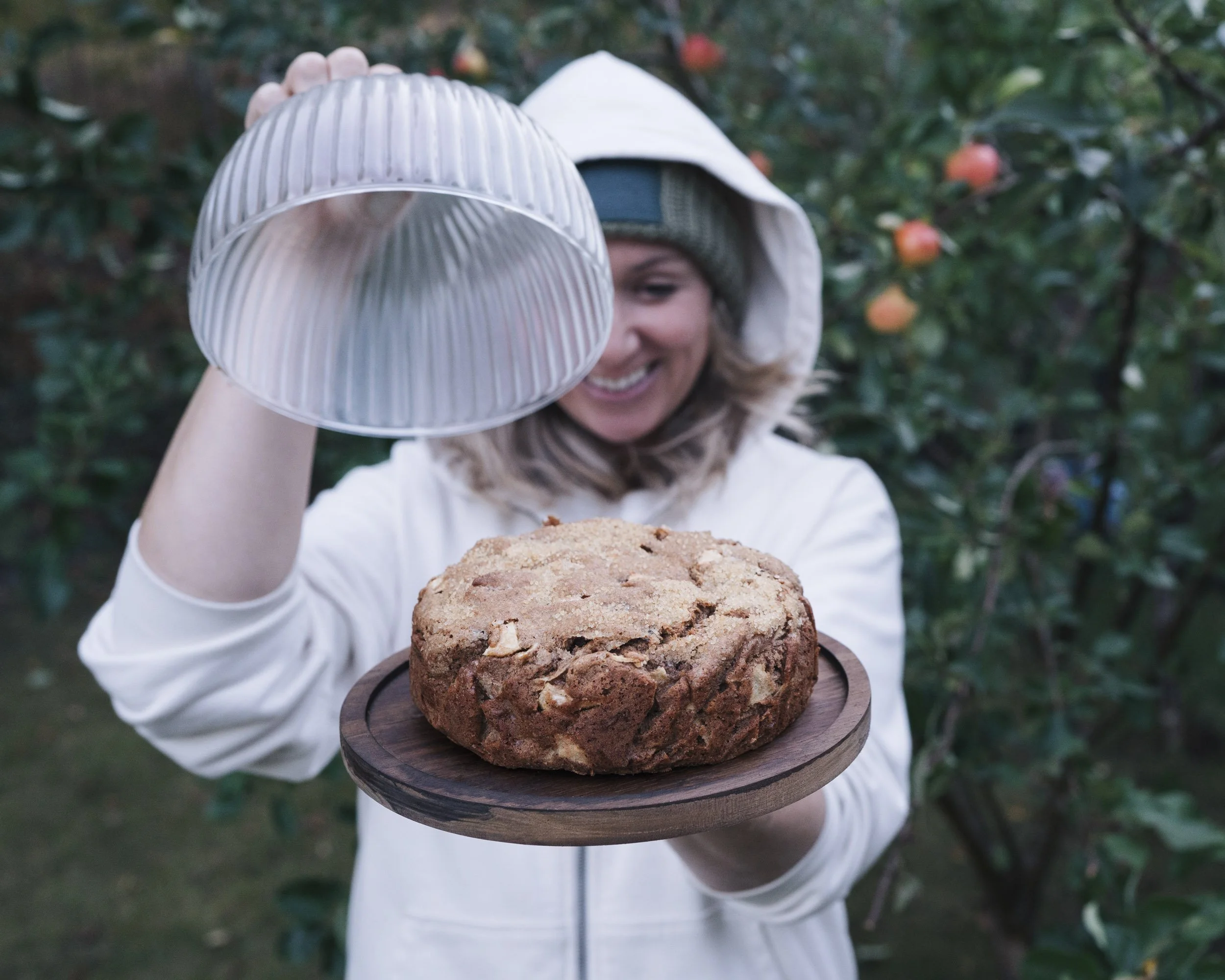 A person wearing a white hoodie and a gray beanie, holding a wooden tray with a large chocolate chip cookie, with trees and bight orange fruits in the background.