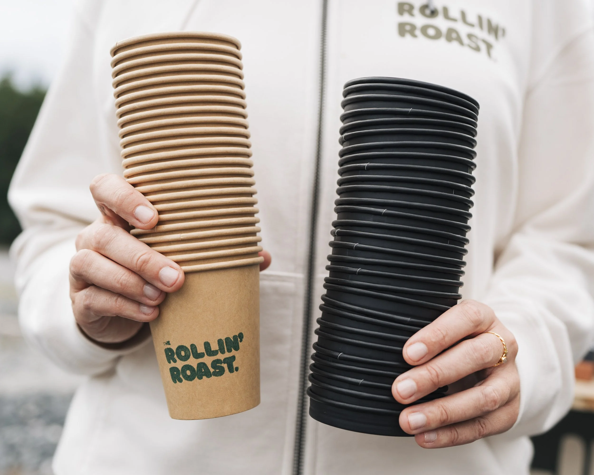 Person holding a stack of tan disposable coffee cups, and another stack of black disposable coffee cups, both with 'the ROLLIN' ROAST' printed on them, outdoors.