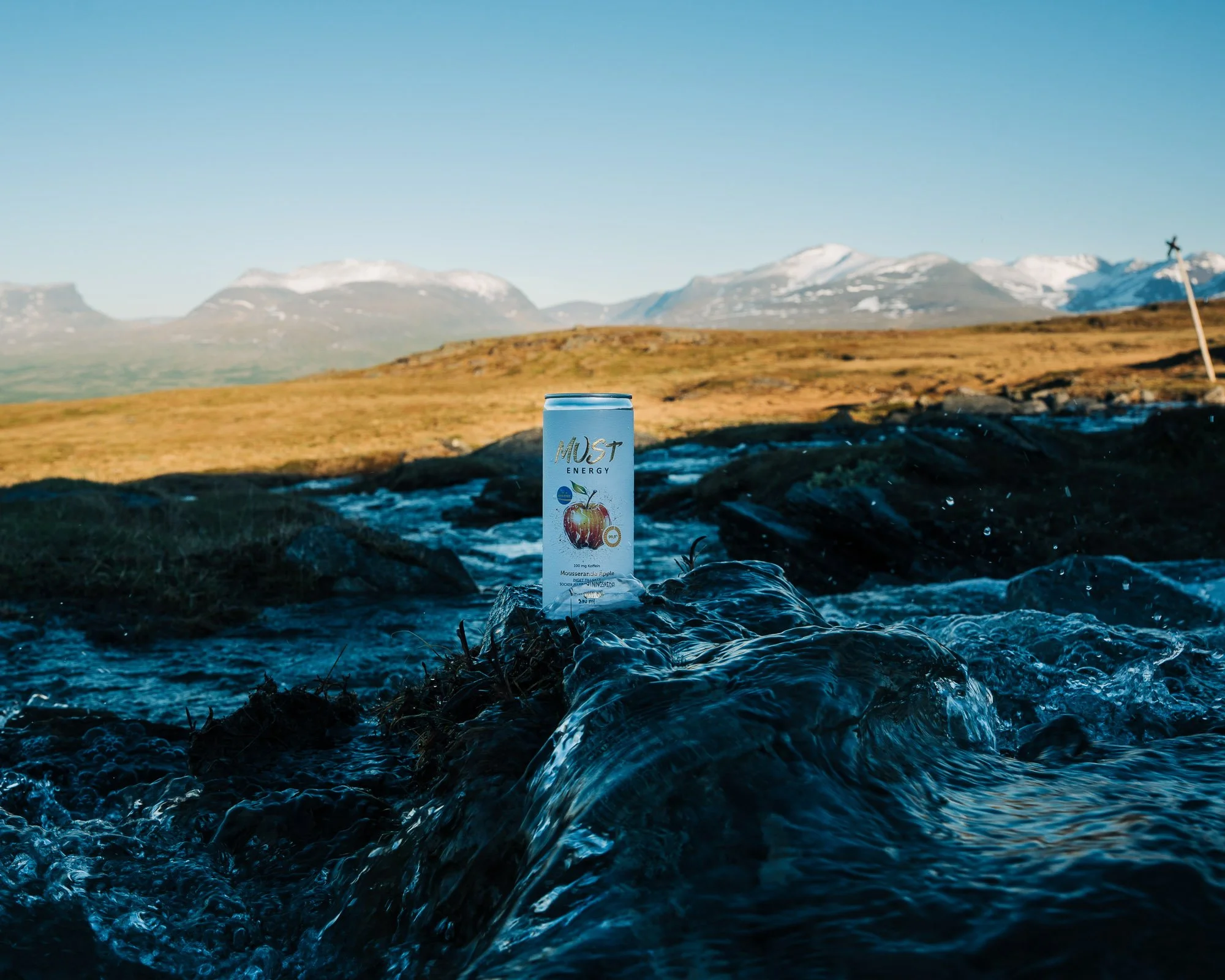 A can of energy drink placed on a rocky surface near a flowing stream, with a mountain range and clear blue sky in the background.