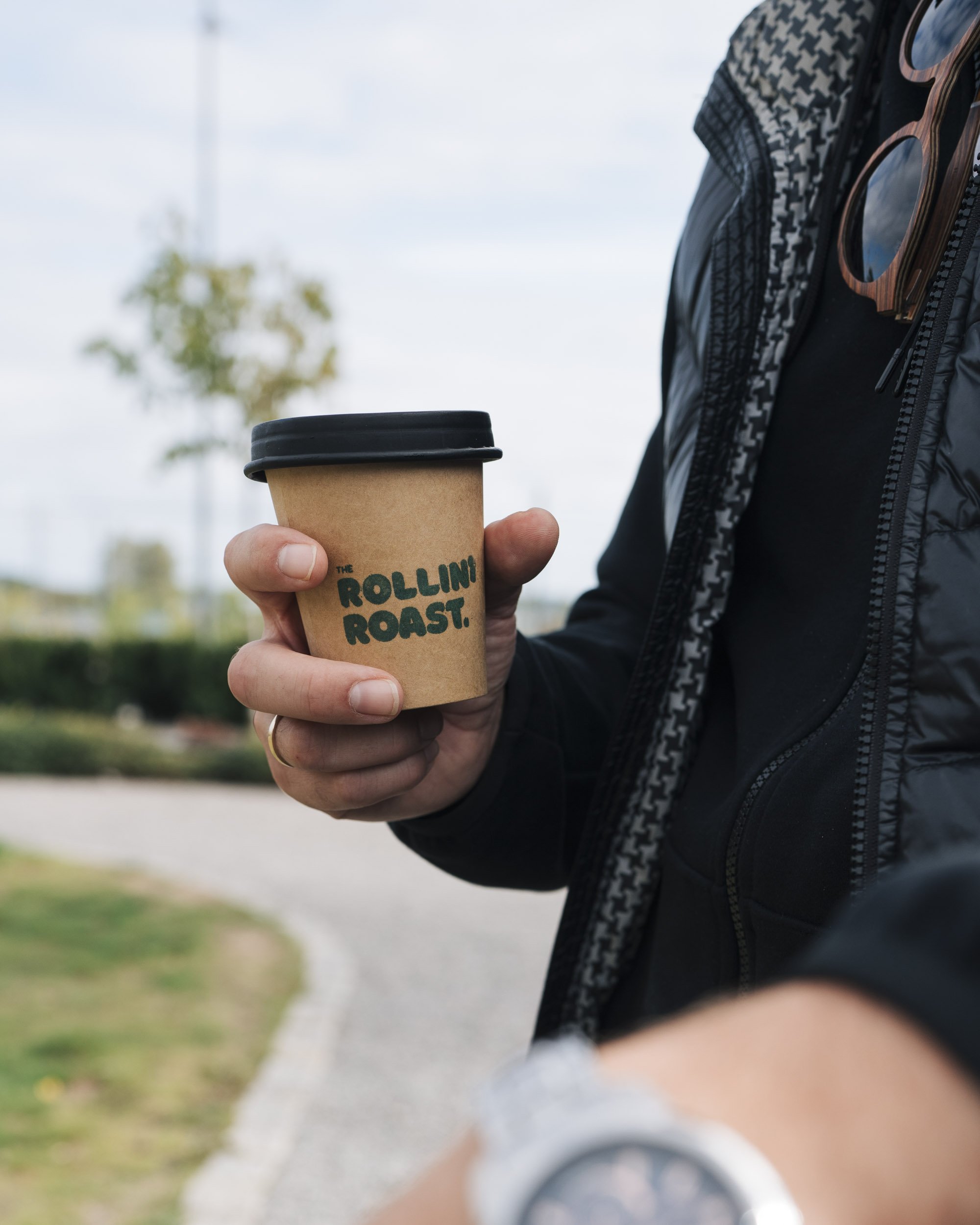 A person in a black jacket holding a brown paper coffee cup with a black lid, with the text 'The Rollin' Roost' on it. The person is outdoors, with blurred trees and sky in the background.