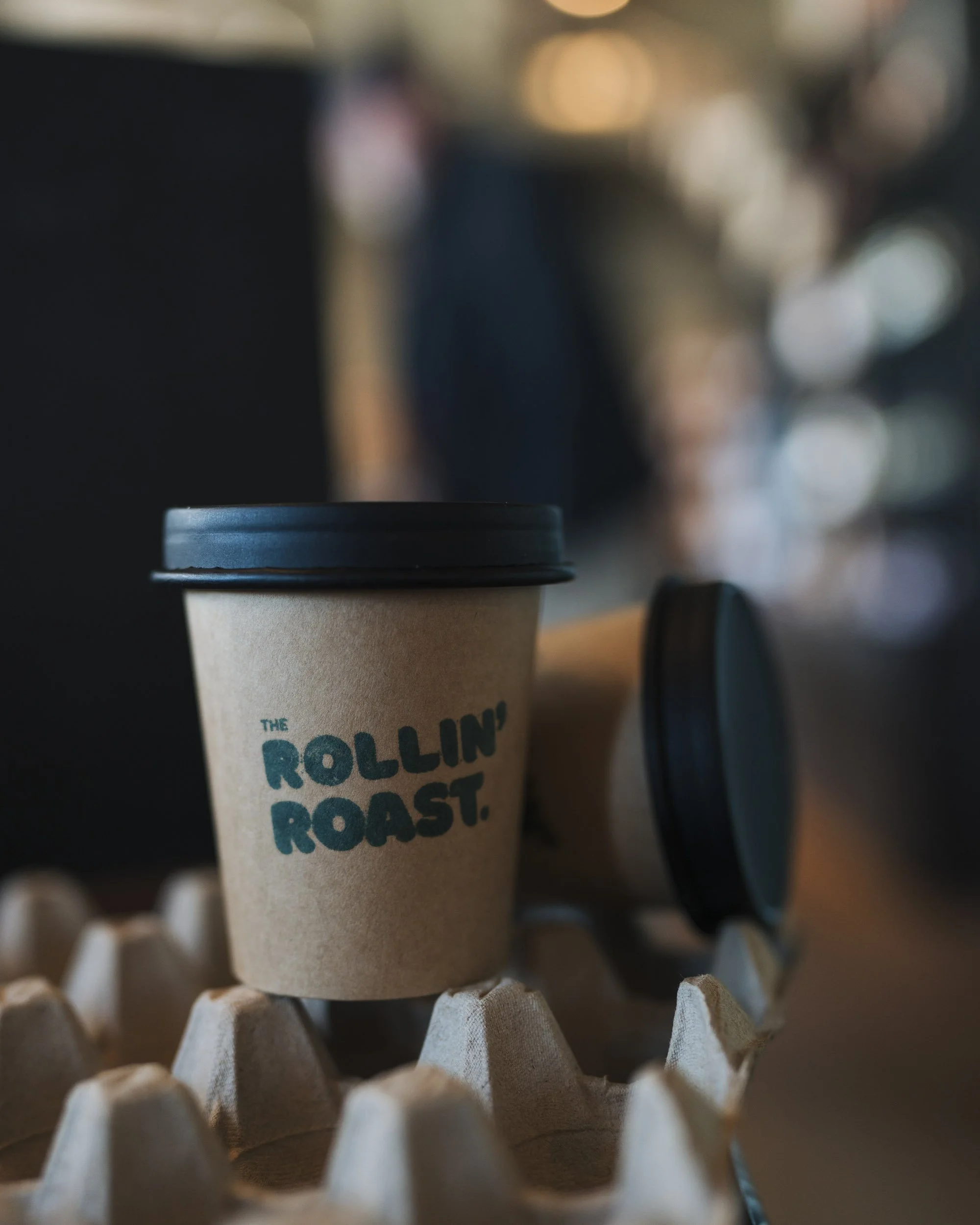 Close-up of a cup of coffee on a paper coffee cup holder, with a blurred background of people and warm lighting in a cafe.