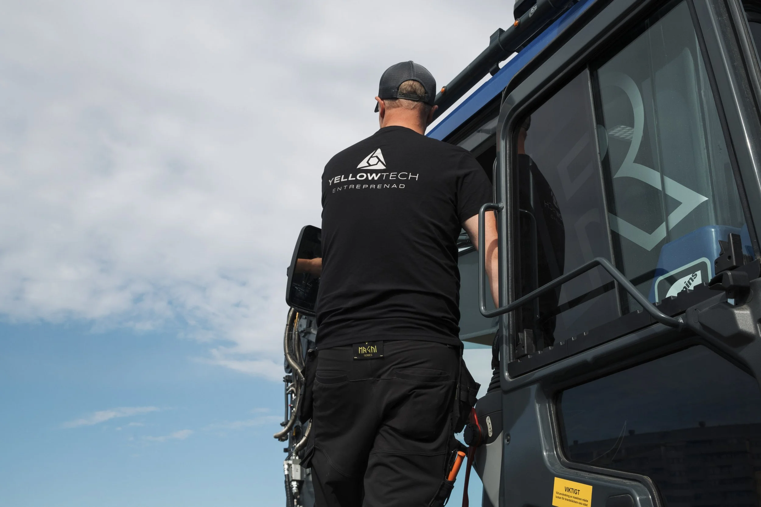 A man wearing a black cap and a black T-shirt with the words 'YELLOWTECH ENTREPRENAD' on the back is operating a large construction vehicle against a cloudy sky background.