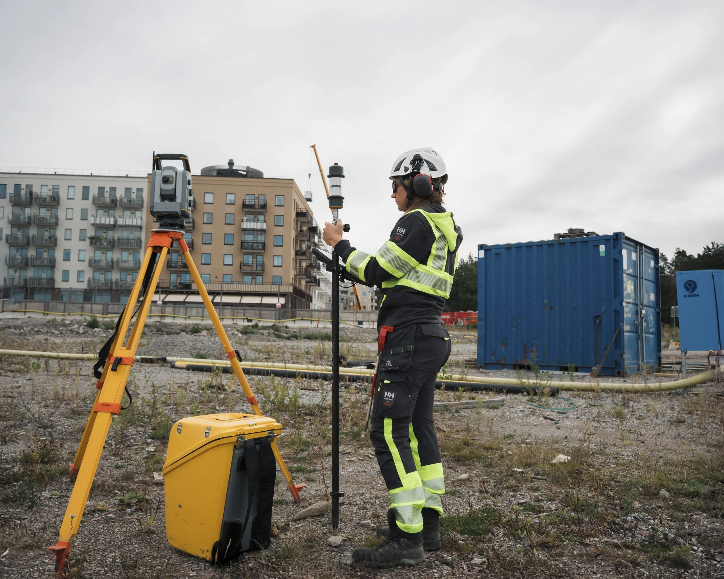 A construction worker in safety gear, including a hard hat and high-visibility clothing, operates surveying equipment on a construction site with apartment buildings in the background under a cloudy sky.