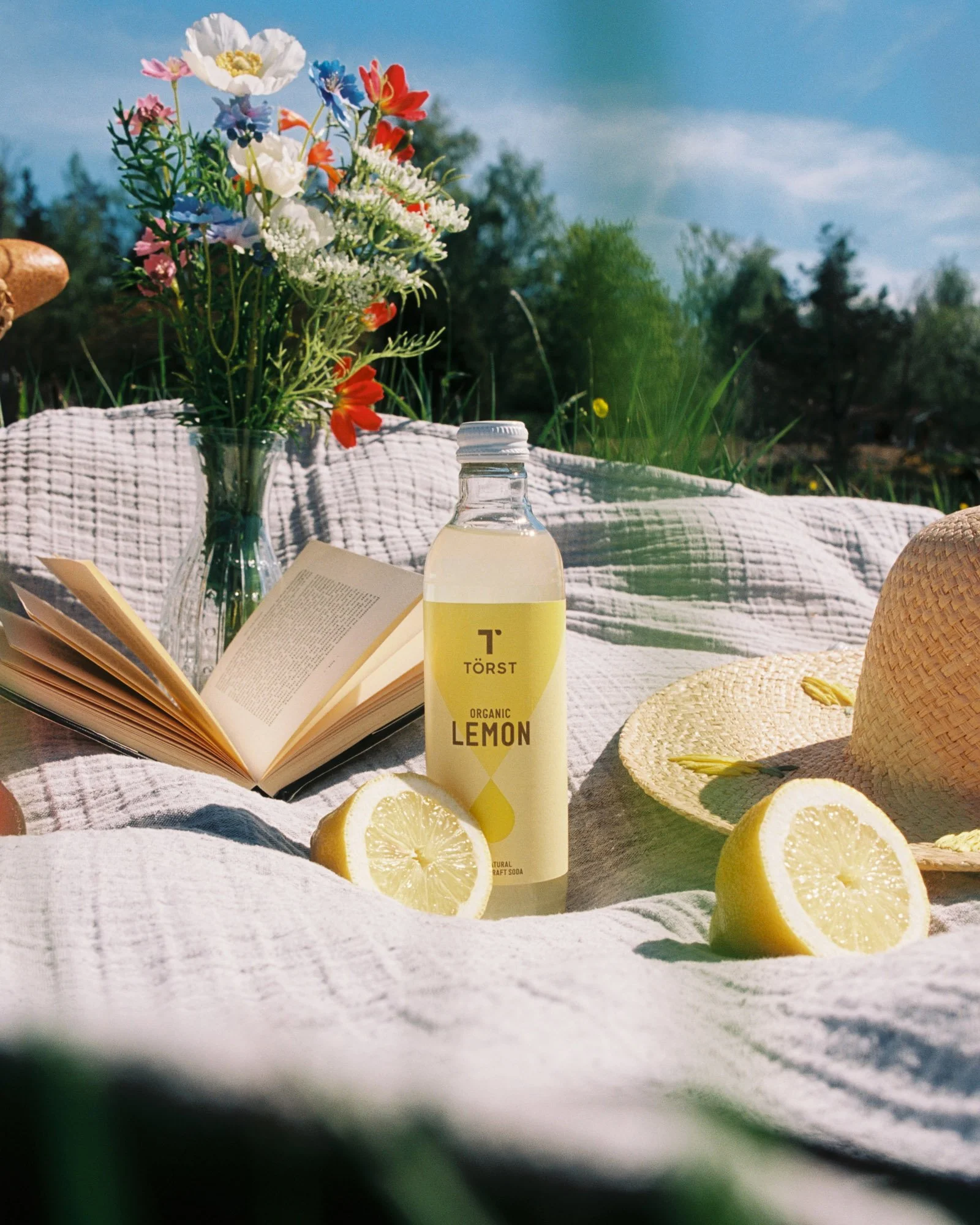A picnic scene with a bouquet of colorful flowers in a clear vase, a yellow bottled organic lemon craft soda, an open book, and lemon slices on a white blanket outside with a background of trees and a blue sky.