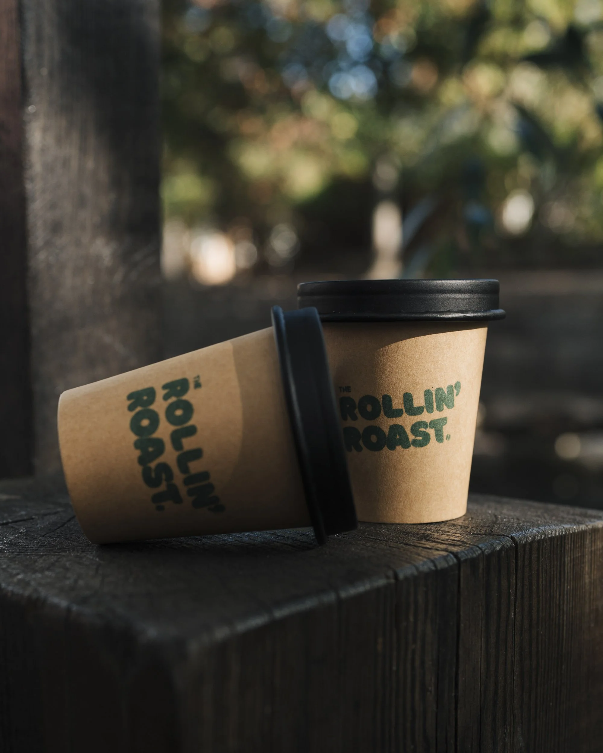Close-up of a coffee cup on a dark wooden surface outdoors, with blurred background of trees and sunlight.