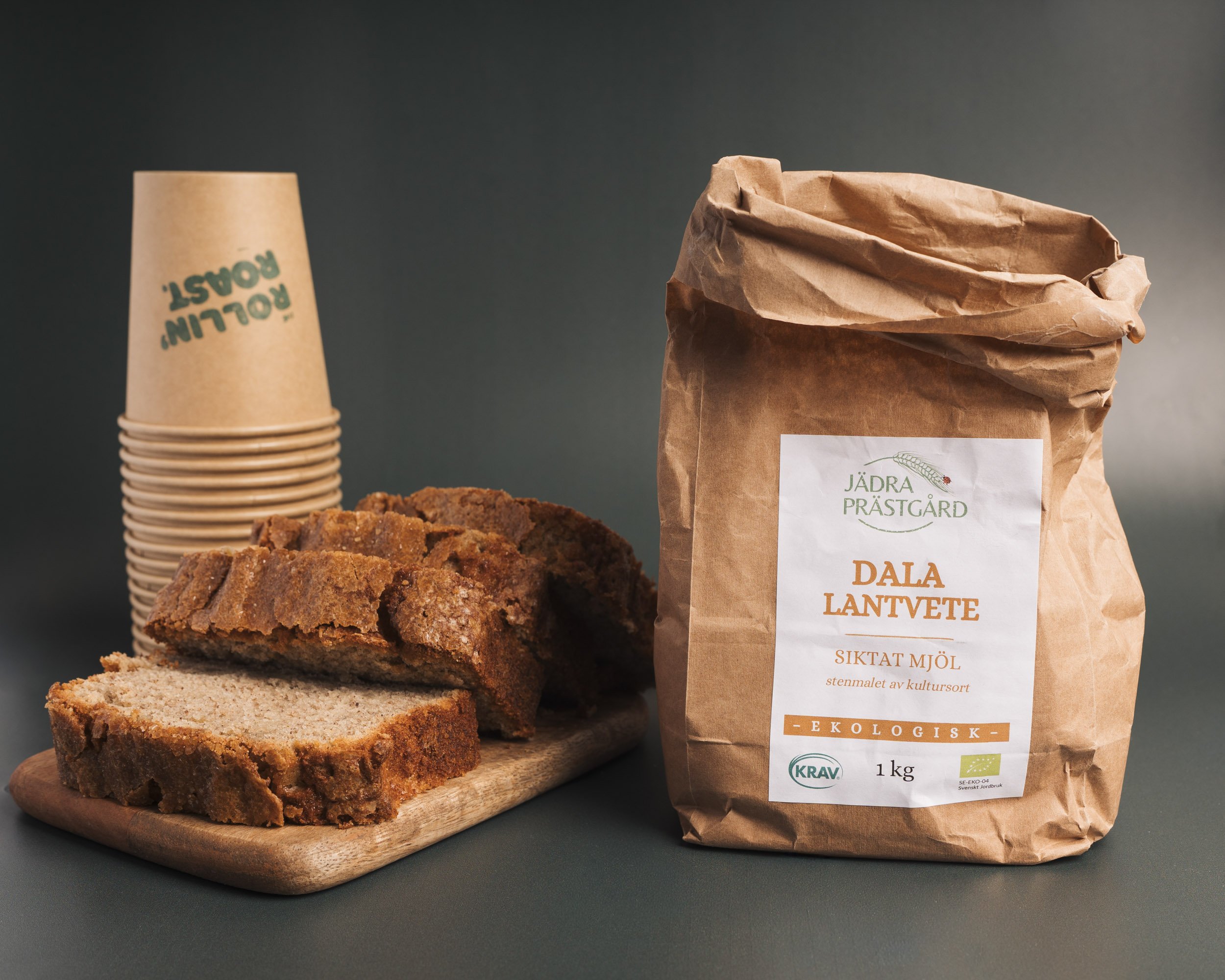 A paper bag of organic, Swedish, farm-grown wheat flour, a loaf of sliced homemade bread, a whole loaf of crusty bread, and a cup of stacked disposable cups with green text.