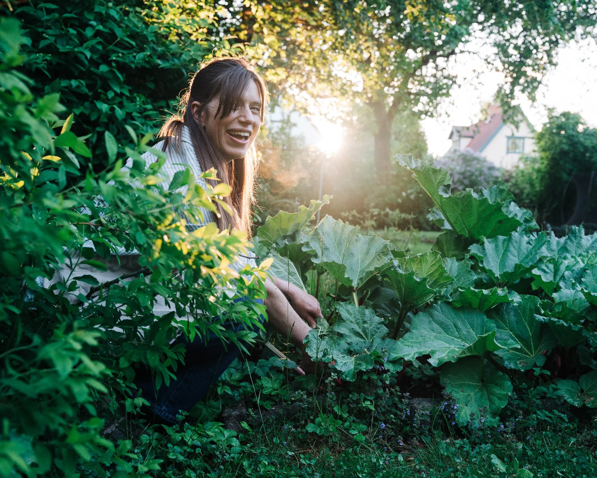 A smiling woman with long brown hair and bangs, wearing a striped shirt, sits among lush green plants and leaves in a garden at sunset.