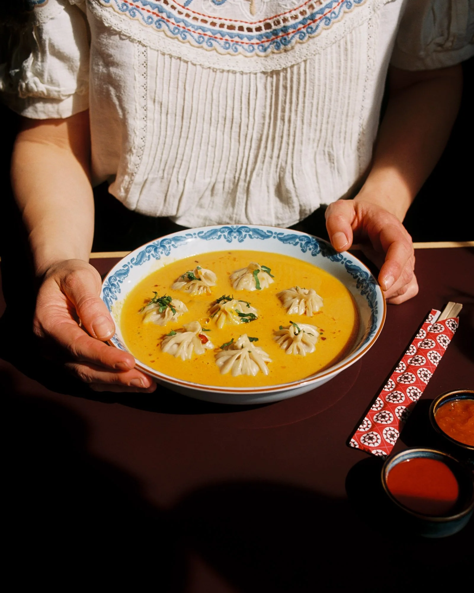 A person in a white embroidered blouse holding a bowl of yellow curry with dumplings and herbs, sitting at a table with traditional red and white patterned chopsticks and side dishes.