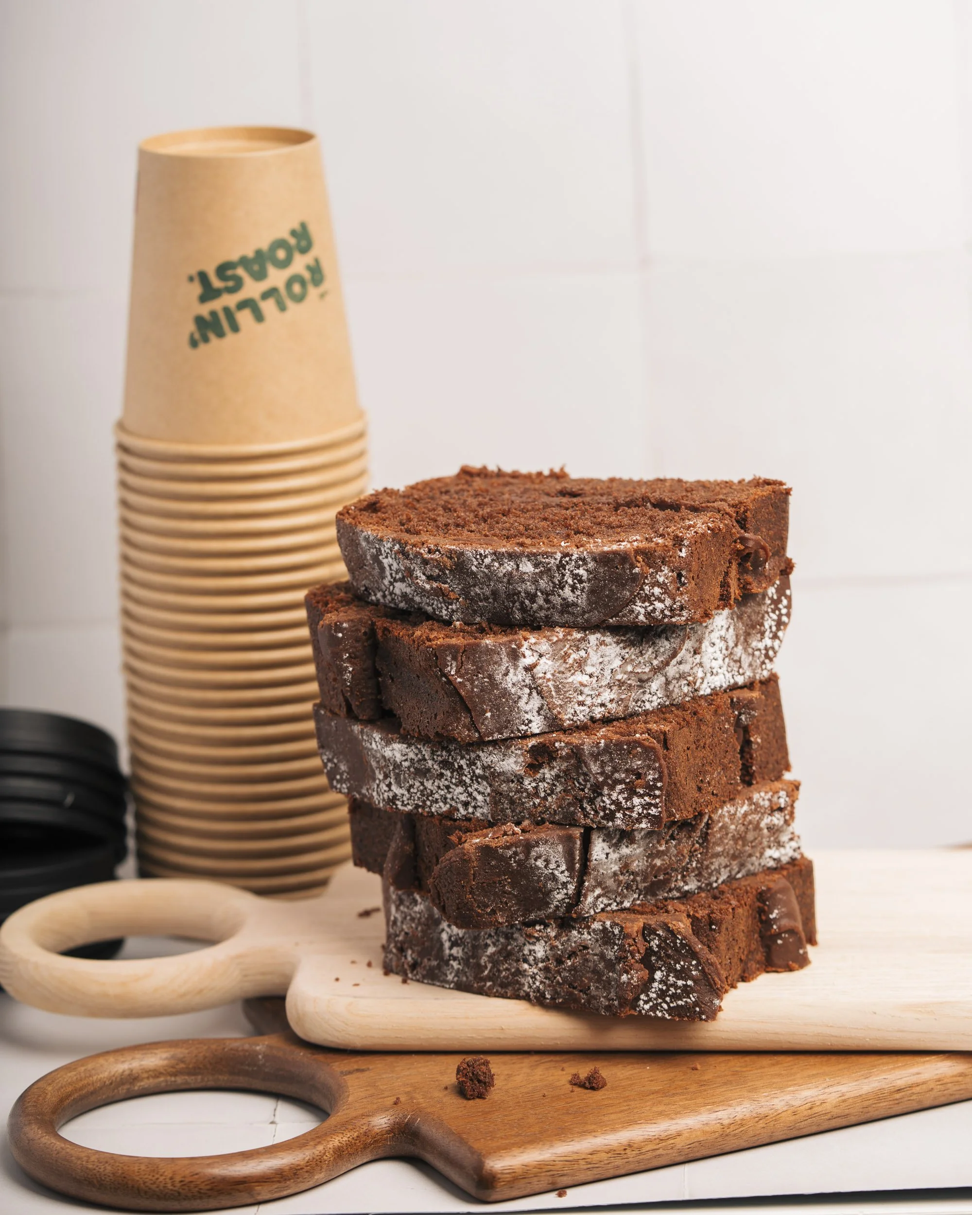 Stacked slices of chocolate cake on a wooden cutting board with a stack of disposable coffee cups in the background.