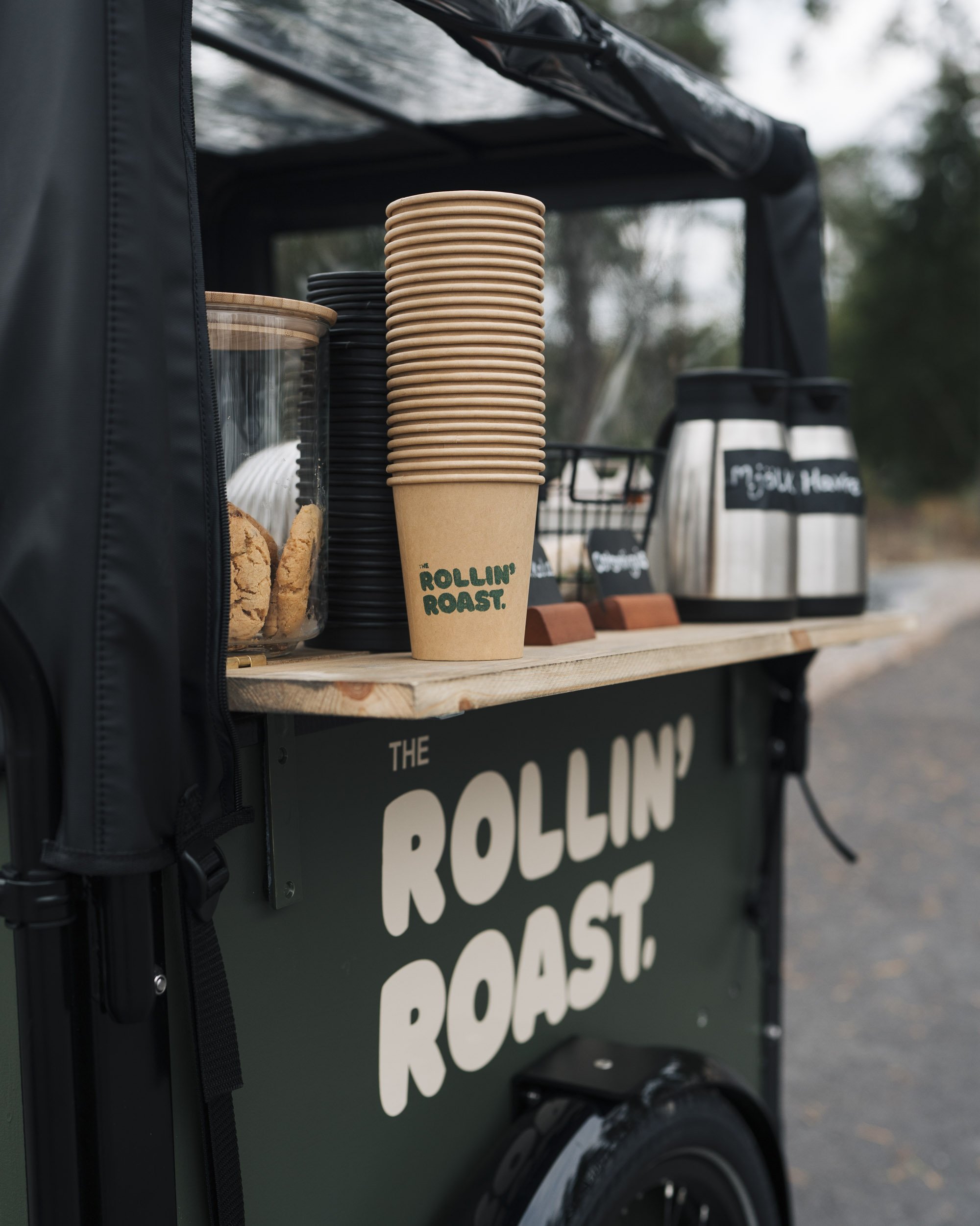 Close-up of a black food cart with a small blackboard sign, stacked paper cups with a green logo reading 'The Rollin' Roast, cookies in a glass jar, stainless steel coffee dispensers labeled 'MELK' and 'COCO' in the background, and a black bag on the
