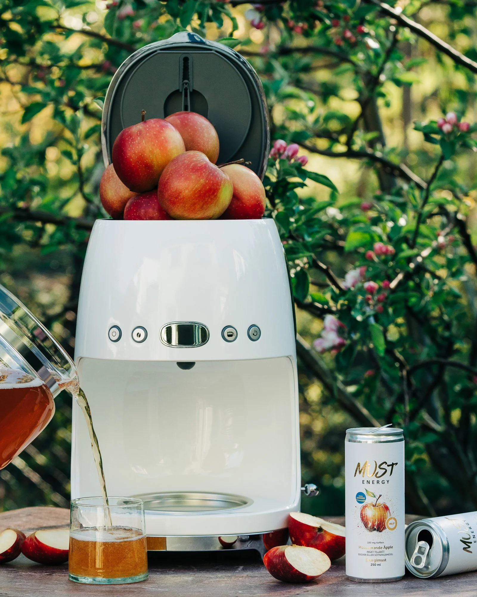 A juicer machine with red apples on top, pouring fresh apple juice into a glass. Surrounding the setup are apples, a can of energy drink labeled "Must Energy," and pink flowering bushes in the background.