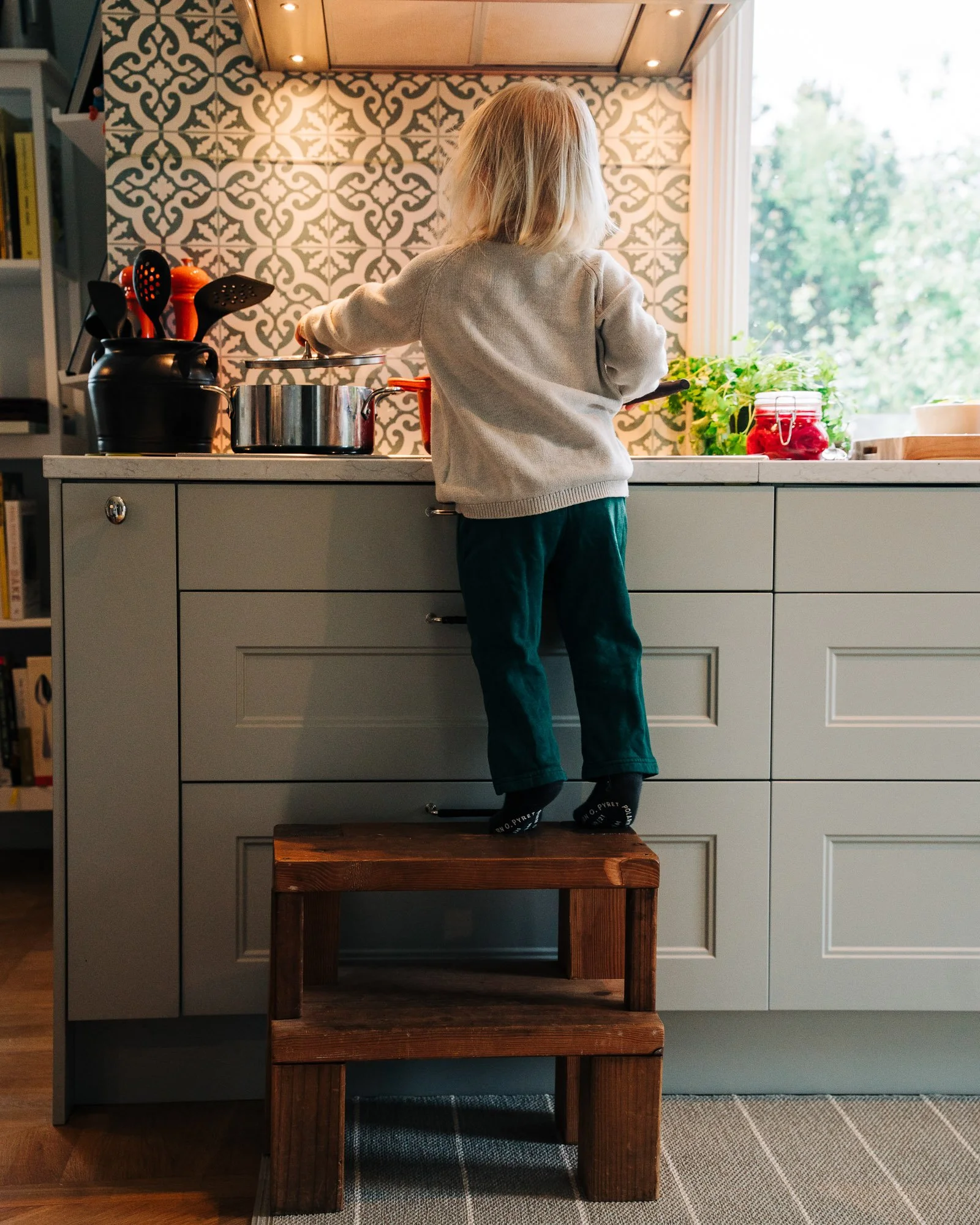 A young child with blonde hair, wearing a beige sweater and dark pants, stands on a wooden stool and reaches toward a kitchen countertop. The kitchen features patterned tiled walls, a window with greenery outside, and various kitchen items including 
