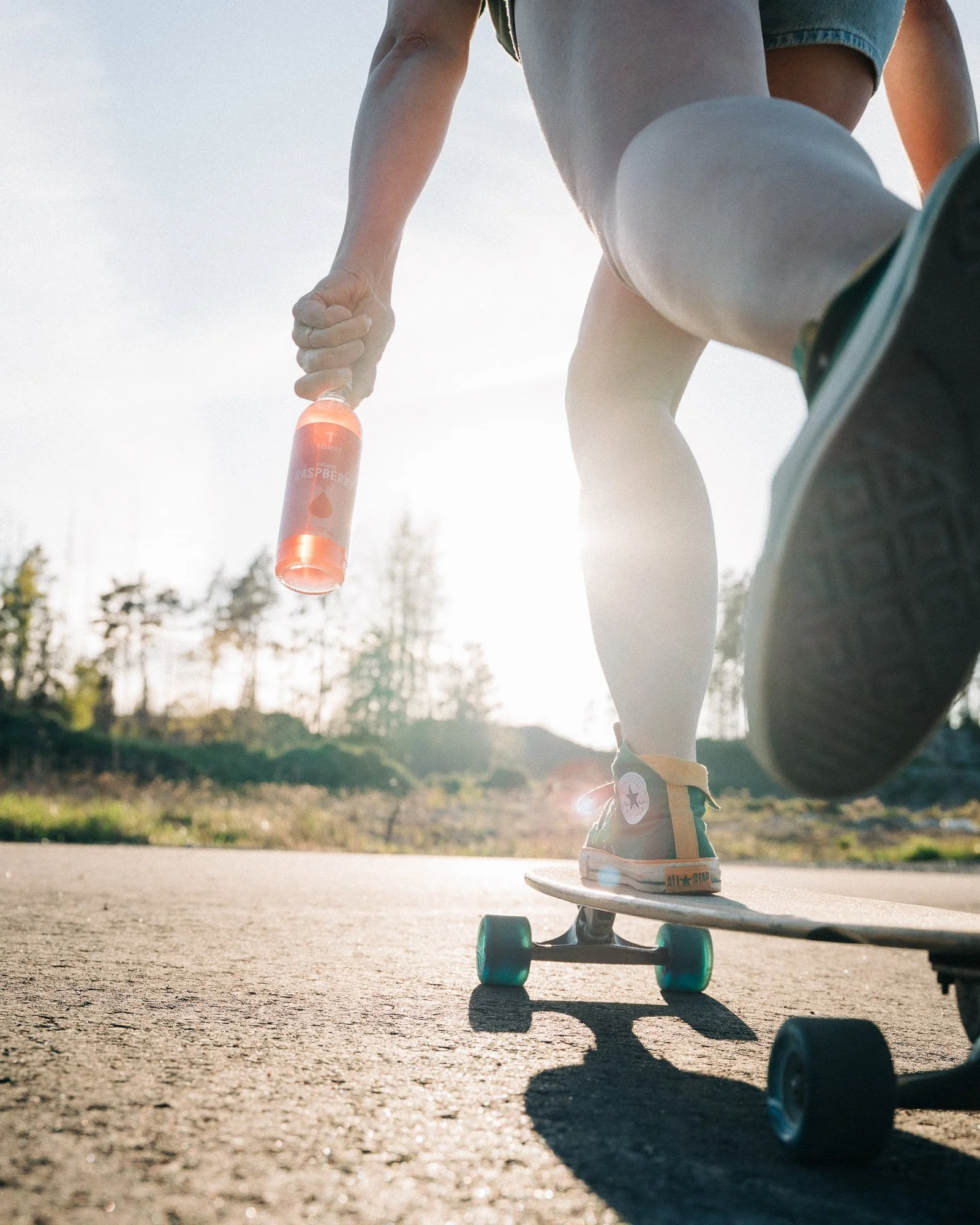 A person performing a trick on a skateboard outdoors on a sunny day, holding a water bottle, with skateboarding shoes and skateboard visible.