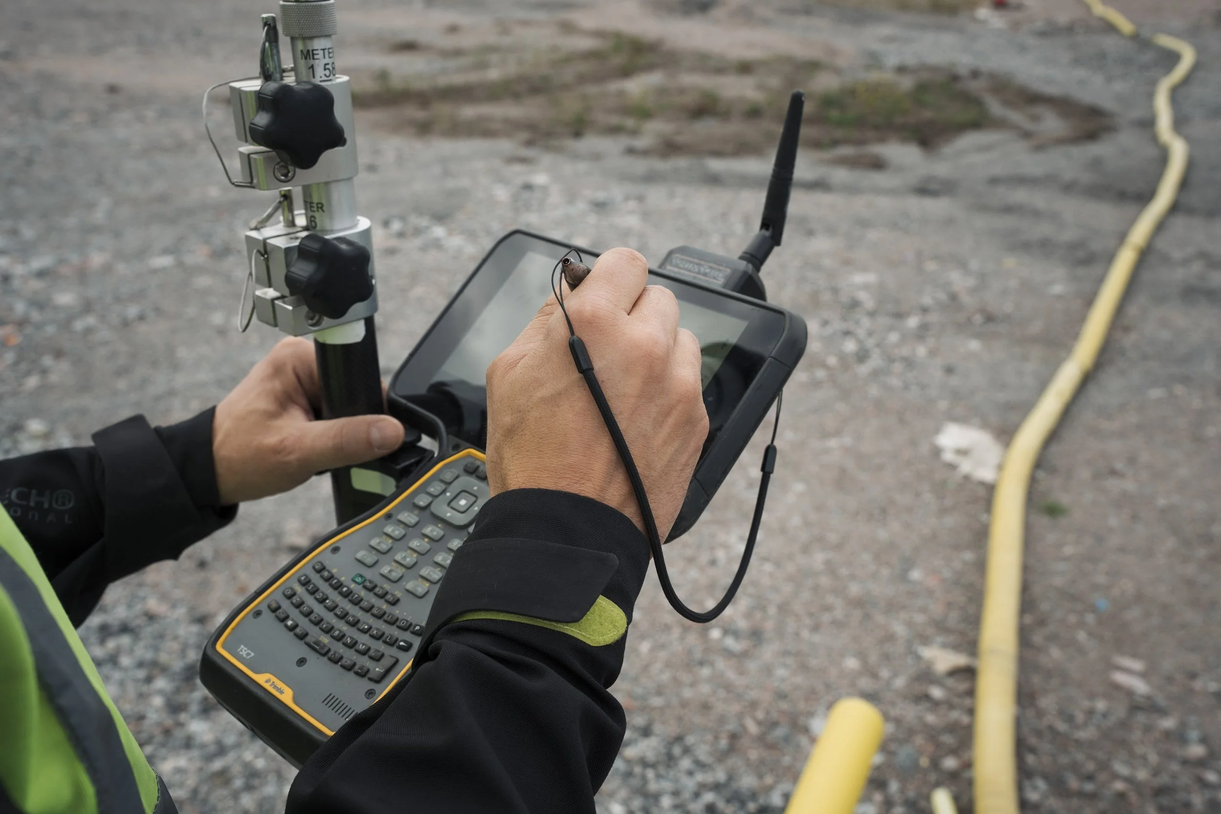 A person using a handheld device and a digital level in an outdoor setting with gravel ground and yellow pipes.