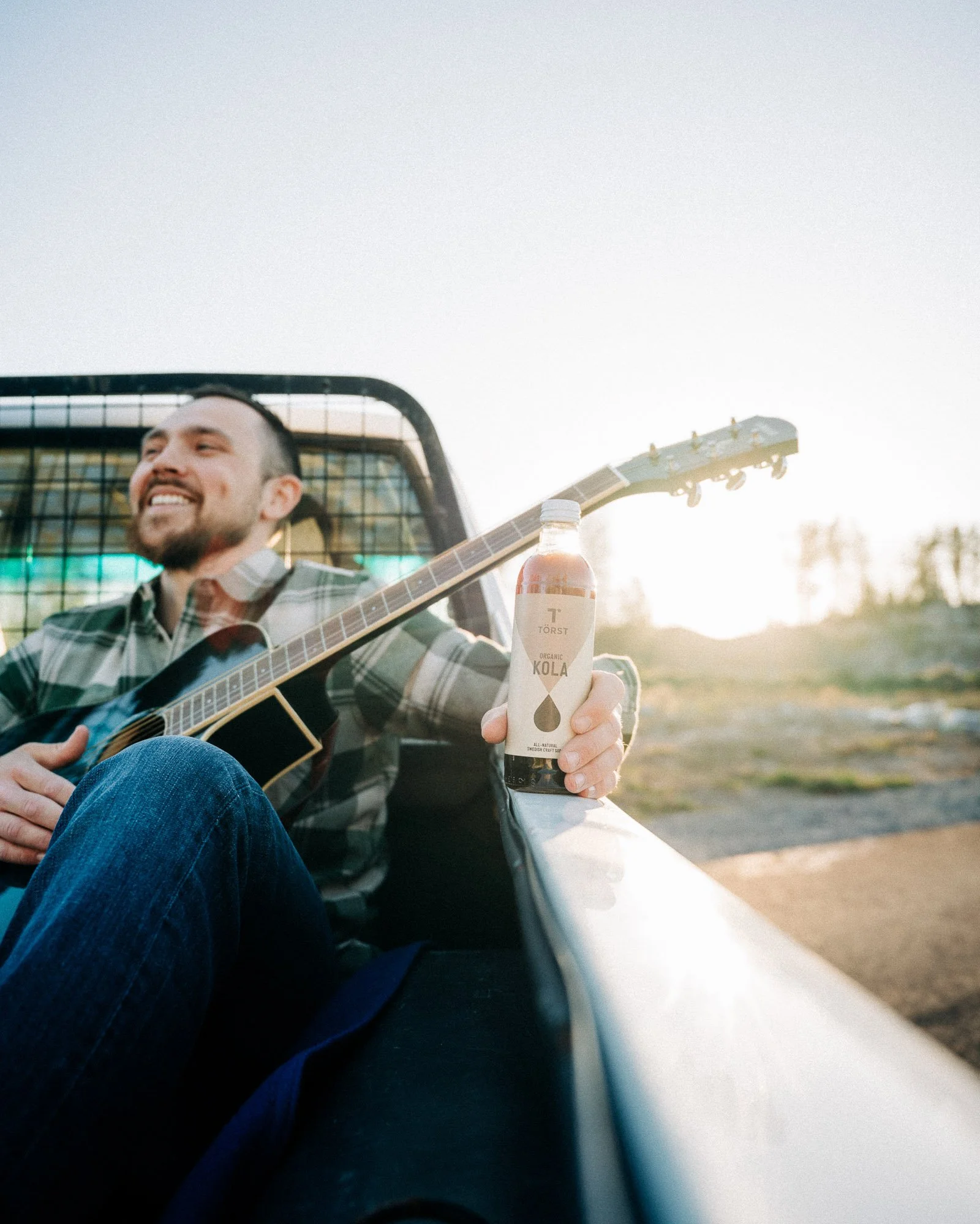A man sitting in a vehicle playing an acoustic guitar and holding a bottle of Törst organic cola, with a sunset in the background.