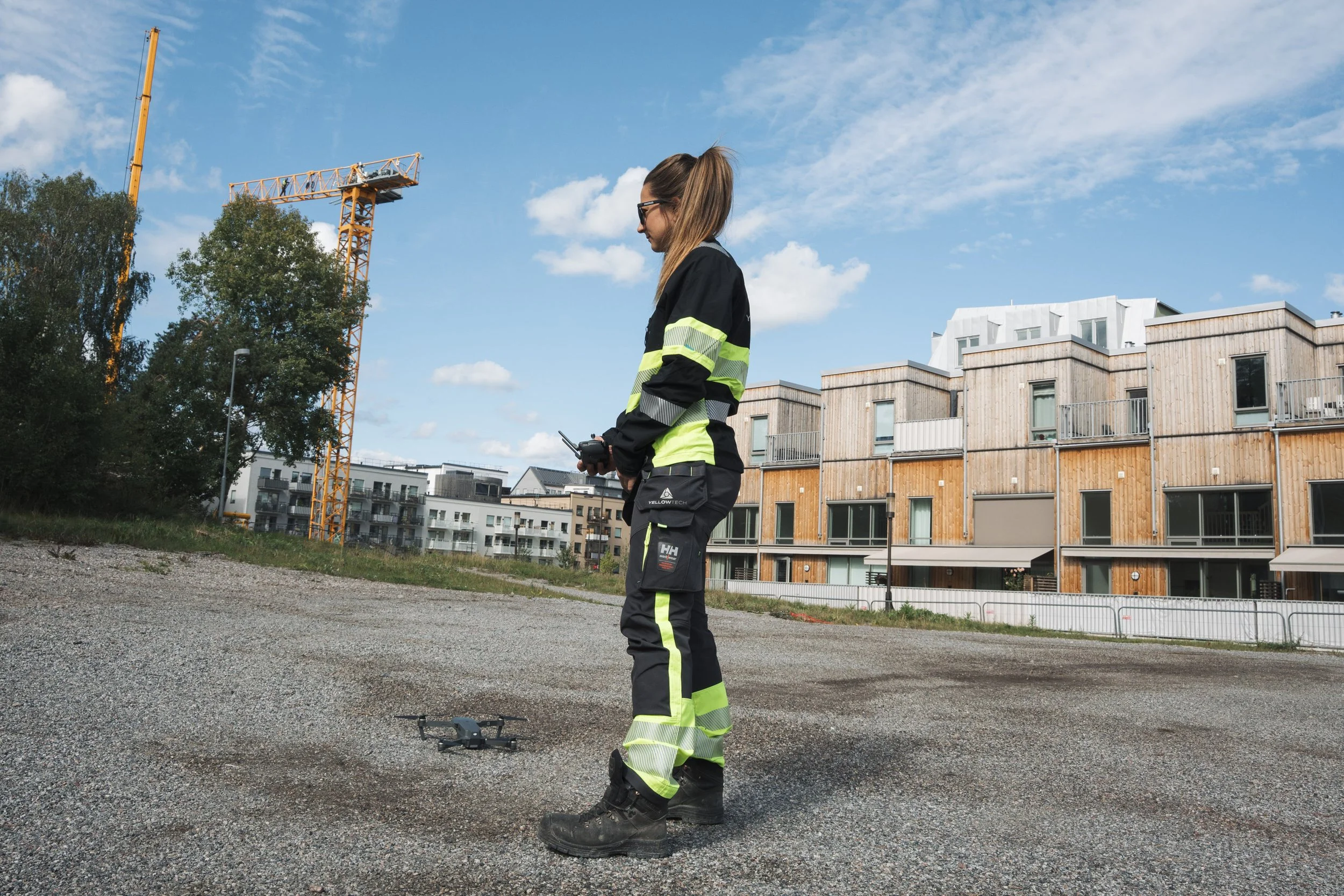 A woman wearing black and neon yellow safety gear using a drone outdoors in a construction area with buildings and a crane in the background.