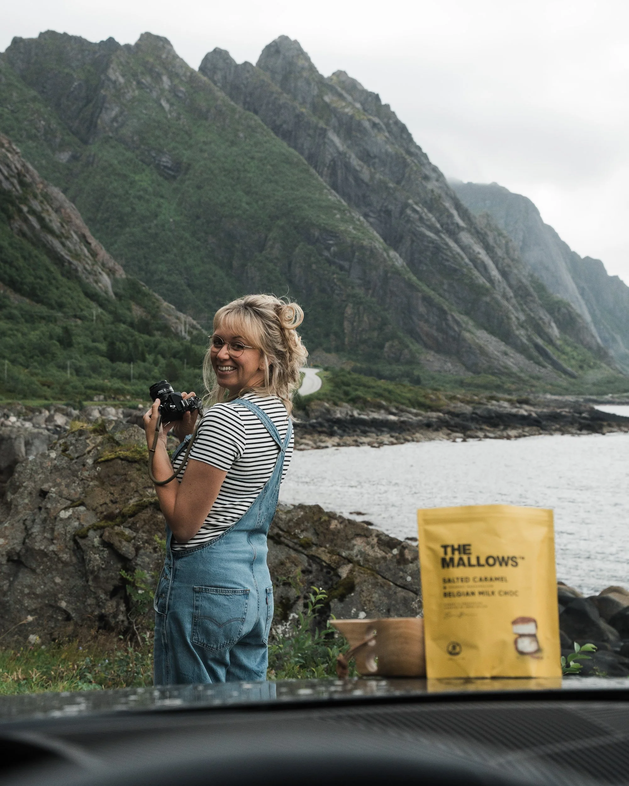 A woman with glasses, blonde hair, wearing a striped shirt and denim apron, smiling and holding a camera, standing outdoors near mountains and a river, with a yellow caramel snack bag on a table in the foreground.