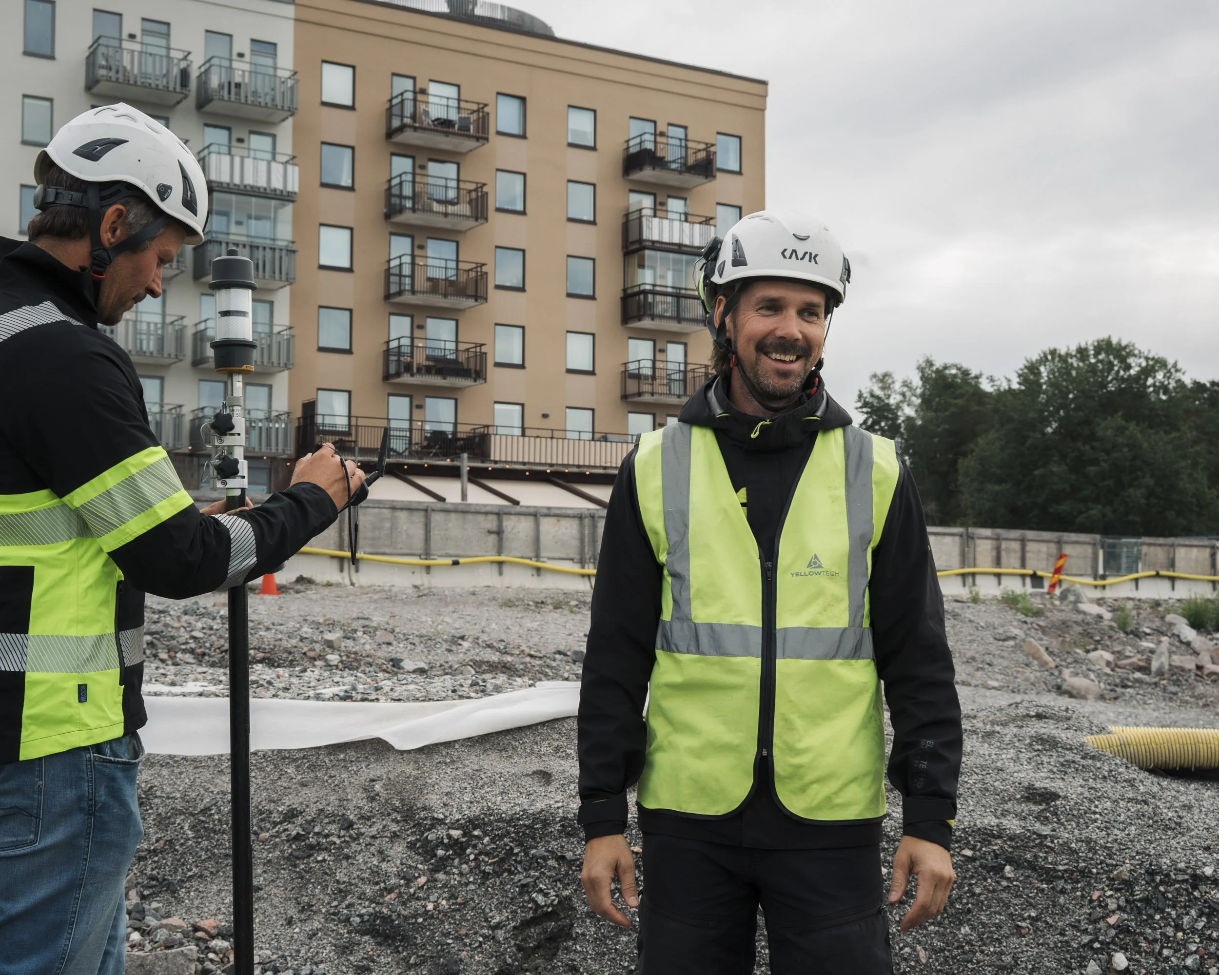 Two men wearing safety helmets and high-visibility vests at a construction site near a residential building. One man is smiling, and the other is looking at equipment. Construction materials and a building with balconies are visible in the background