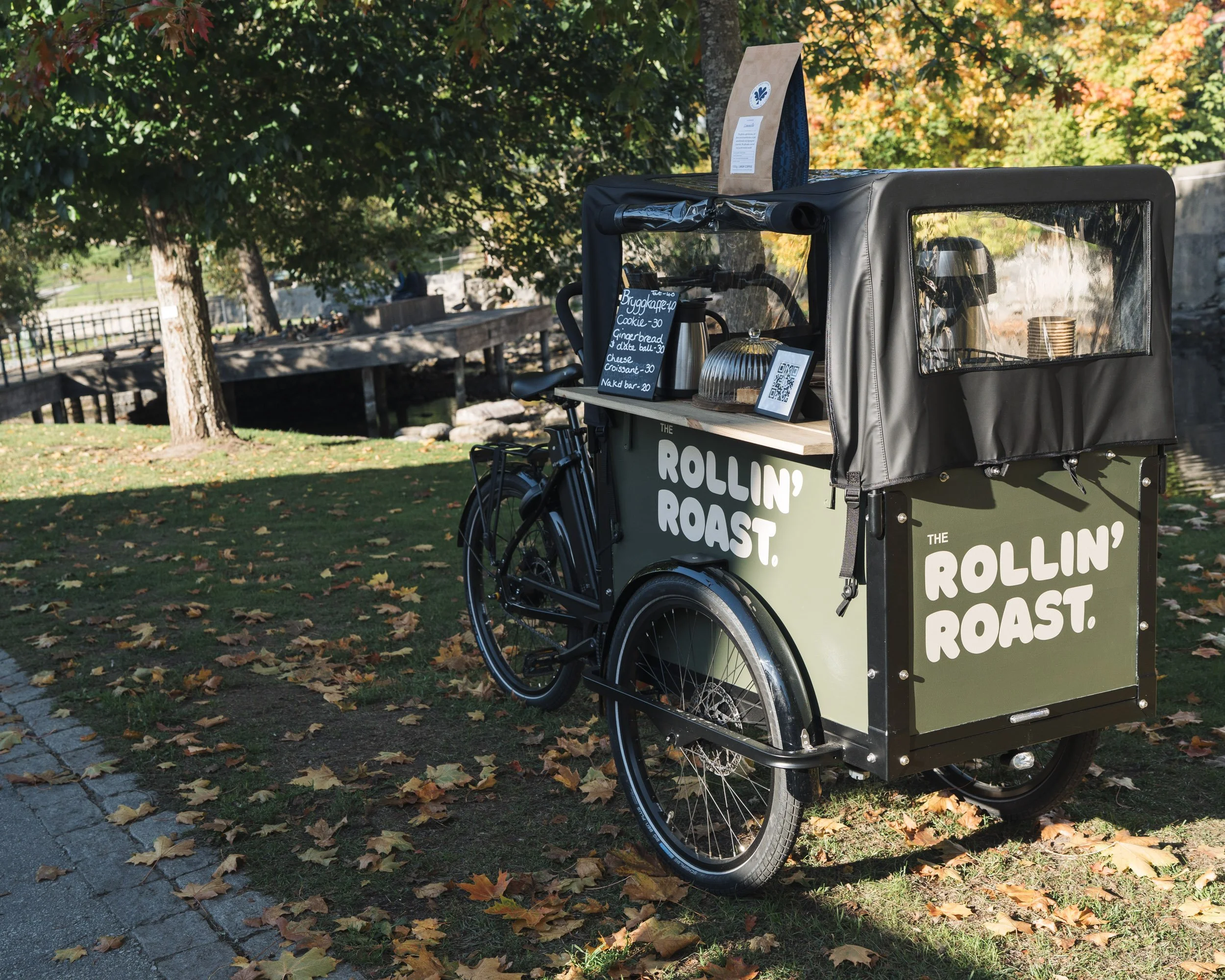 A bicycle with a food cart attached parked on a sidewalk covered with autumn leaves. The cart has a sign on it reading 'The Rollin' Roast' and a menu with items like baked cookies, gingerbread, cheese croissant, and snacks. The background features gr