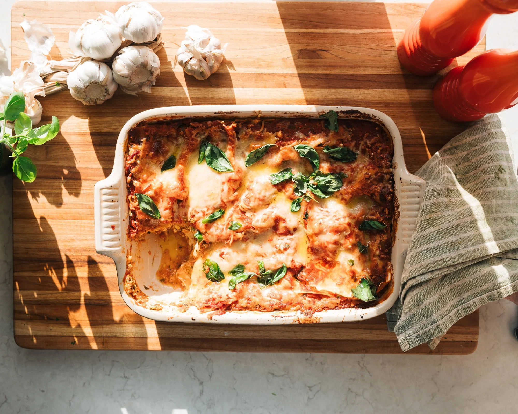 Baked lasagna in a white dish garnished with basil leaves, placed on a wooden cutting board with garlic bulbs and hot sauce bottles nearby, and a striped cloth on the side.