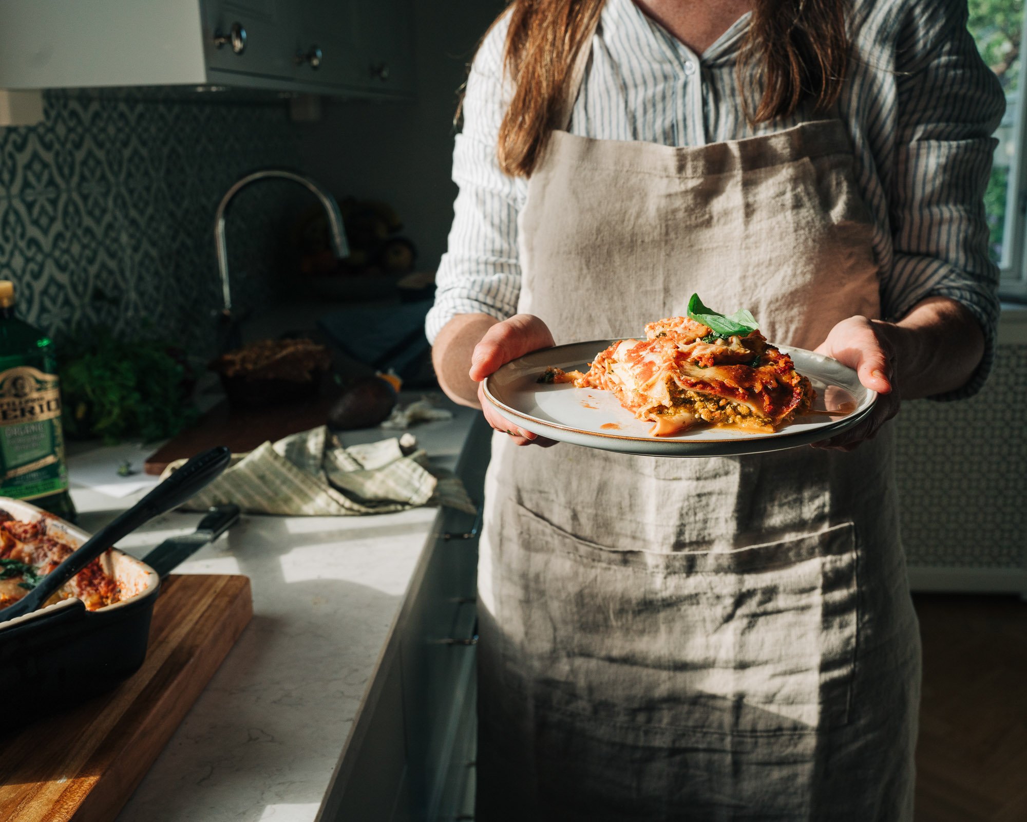Person holding a plate of lasagna in a kitchen with cooking ingredients on the counter.