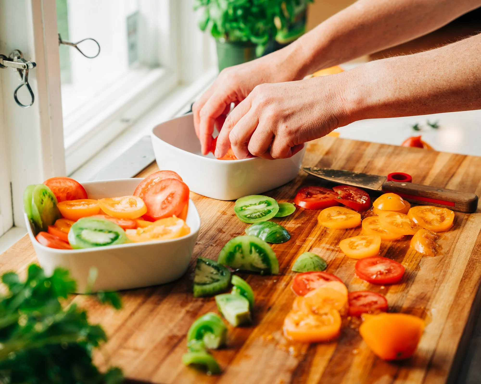 Person placing sliced tomatoes into a white bowl on a kitchen counter with other vegetables on a cutting board.
