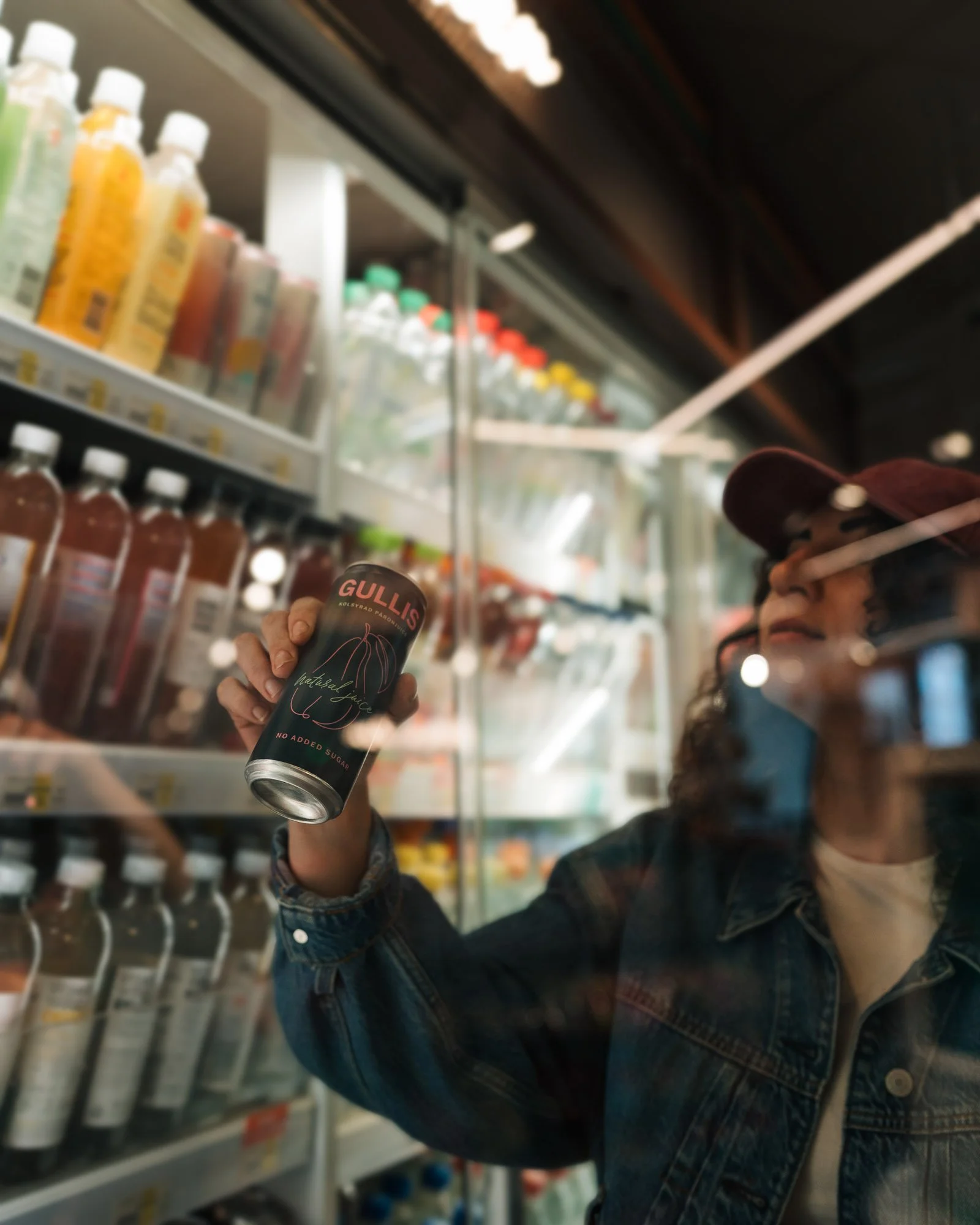 A person holding a can of GULLIS natural juice with no added sugar in a grocery store aisle. The store shelves are stocked with bottles of various beverages, and the person is wearing a denim jacket and a baseball cap.