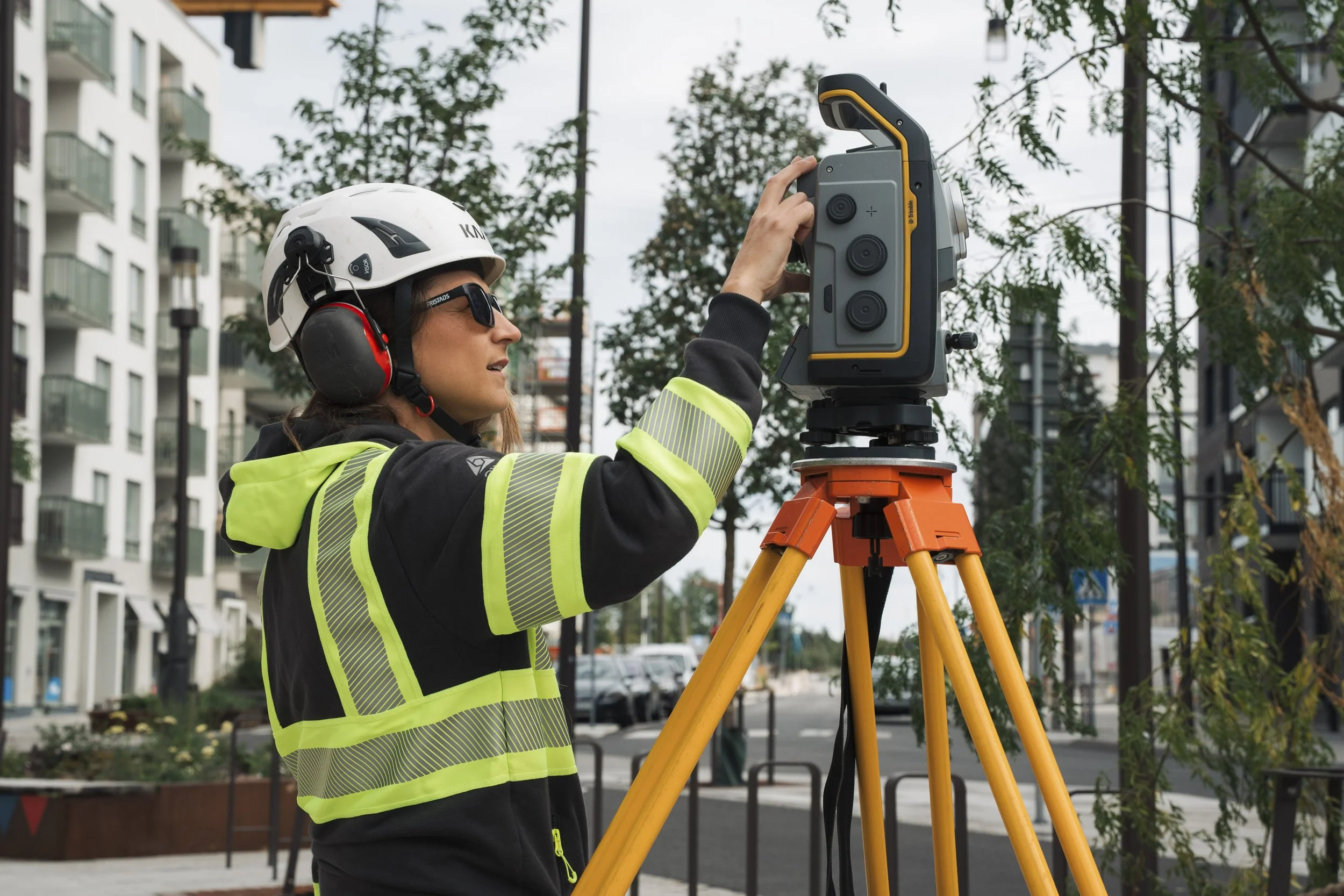 A woman wearing safety glasses, a white helmet, and a high-visibility jacket is operating a surveying instrument mounted on a tripod outdoors, with trees and buildings in the background.