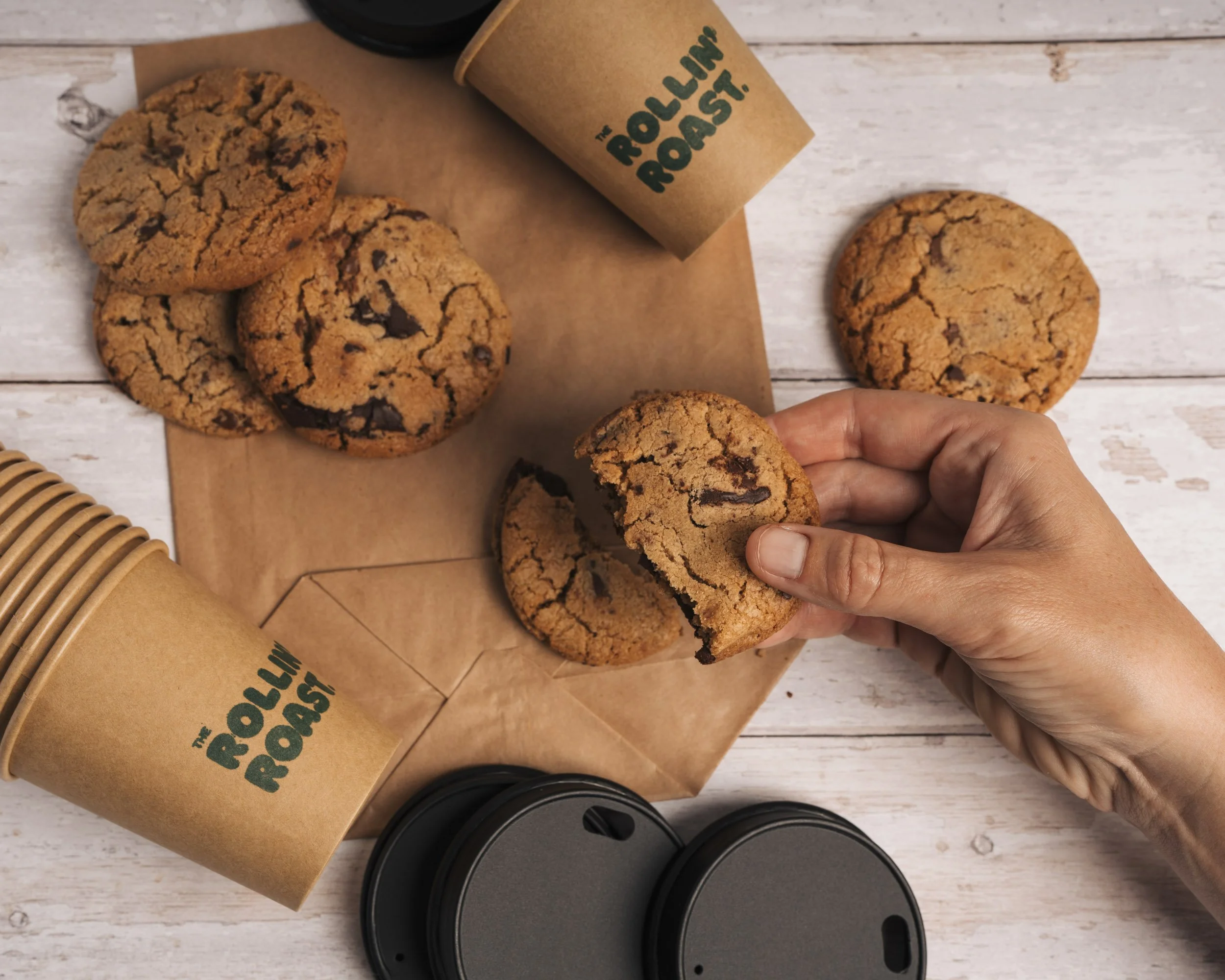 Three freshly baked chocolate chip cookies on a wooden surface, one being held by a person's hand, with coffee cups and coffee lids nearby, and a paper bag labeled 'The Rolling Roost'.