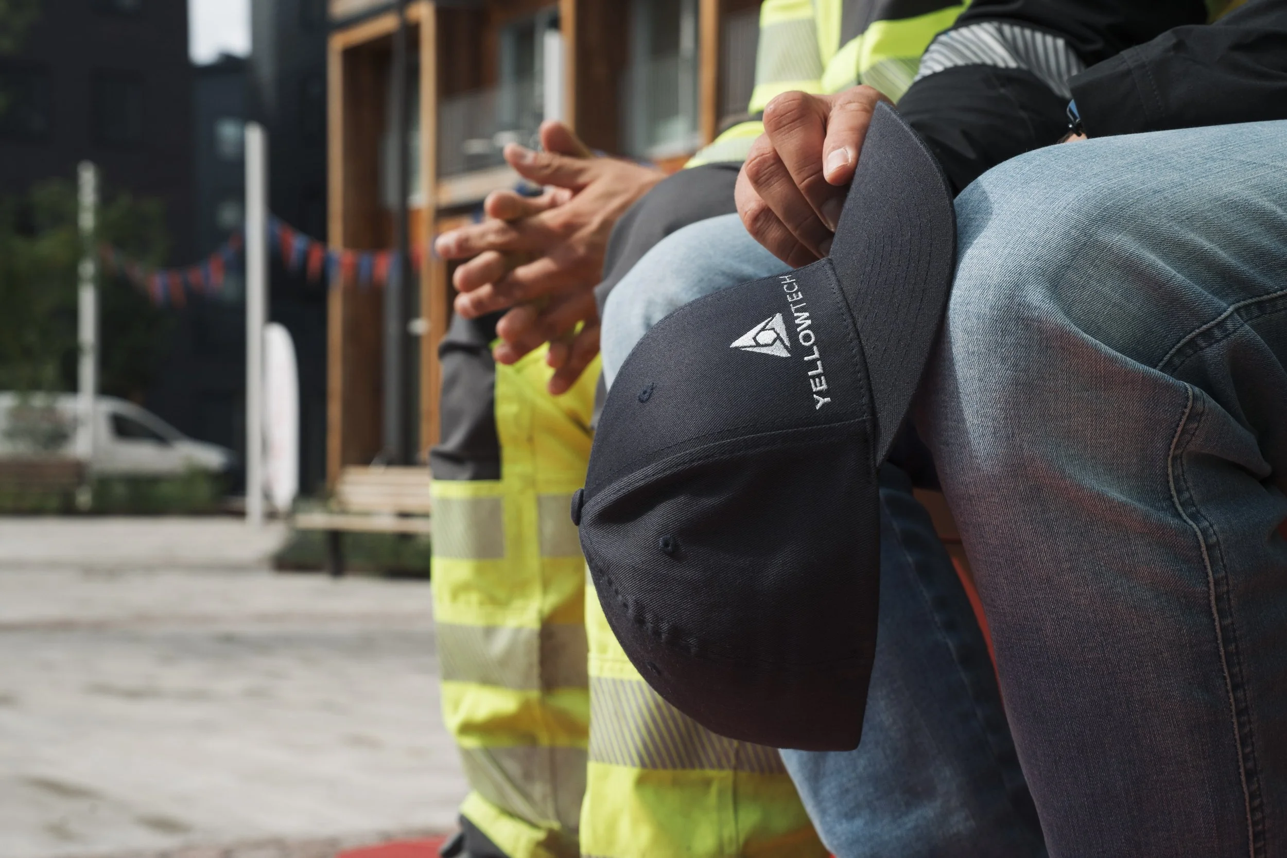 People sitting on a bench, some wearing high-visibility vests, with one person holding a black work cap with the 'YELLOWTECH' logo visibly displayed.