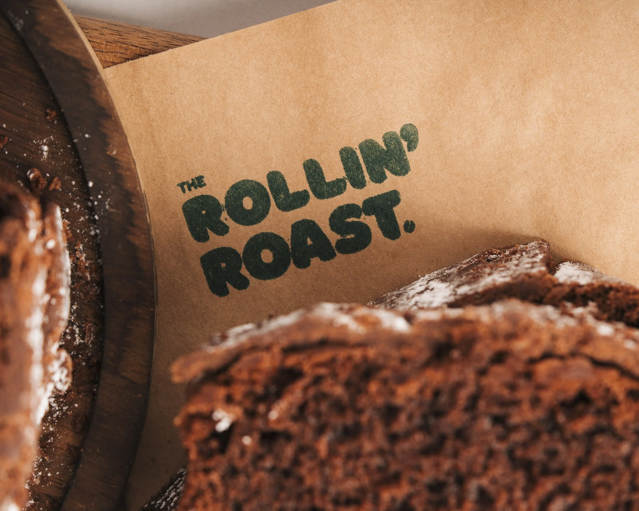 Close-up of a brown paper food packaging with the words 'The Rolling Roast' printed in green, placed amongst baked or roasted breadsticks or bread slices with a dark crust.