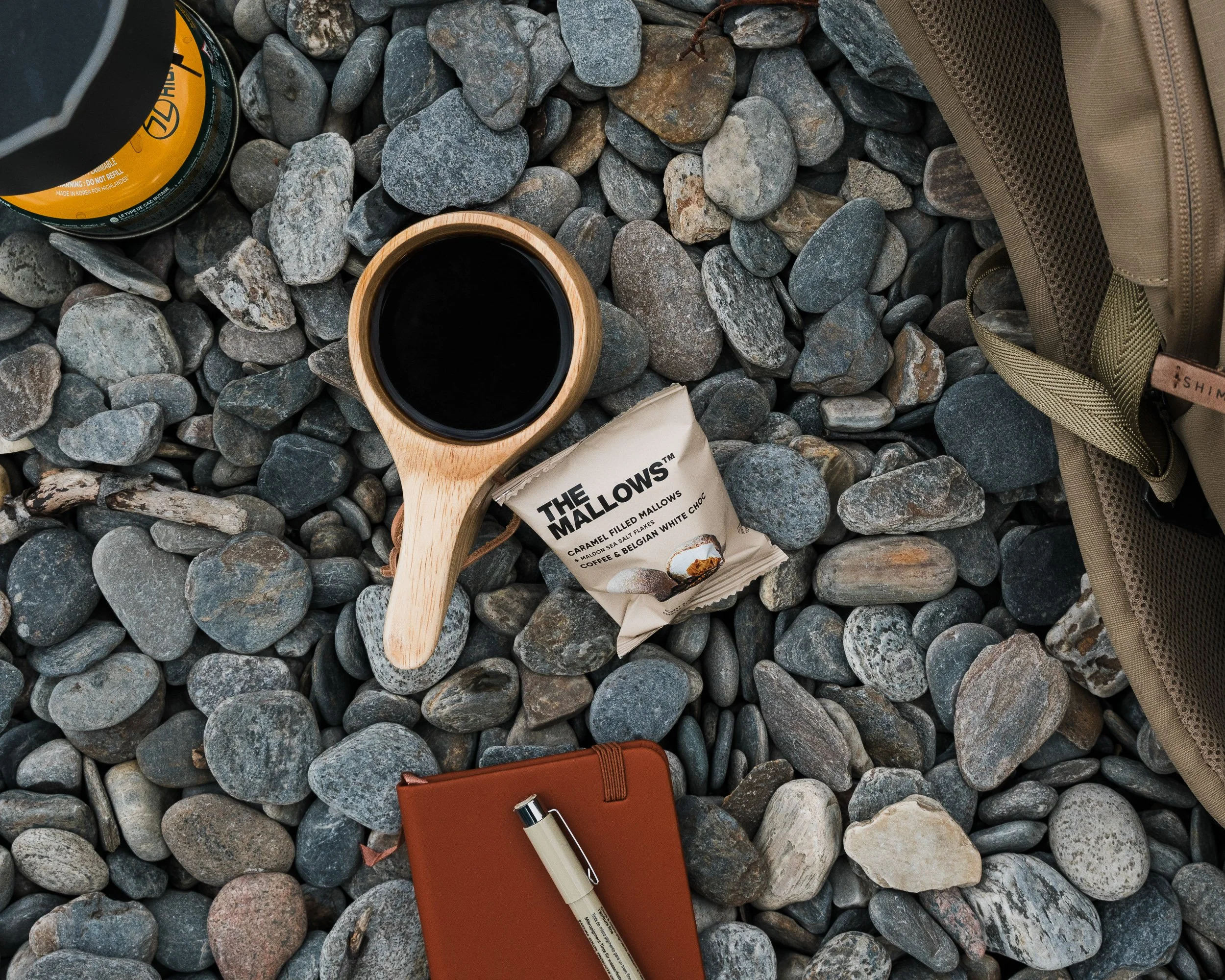 A flat lay of various items on gray rocks, including a yellow-capped can, a cup of black coffee in a wooden cup, a beige backpack, a white pen, a brown notebook, a packet of caramel-filled mallows candies, and additional smaller objects.