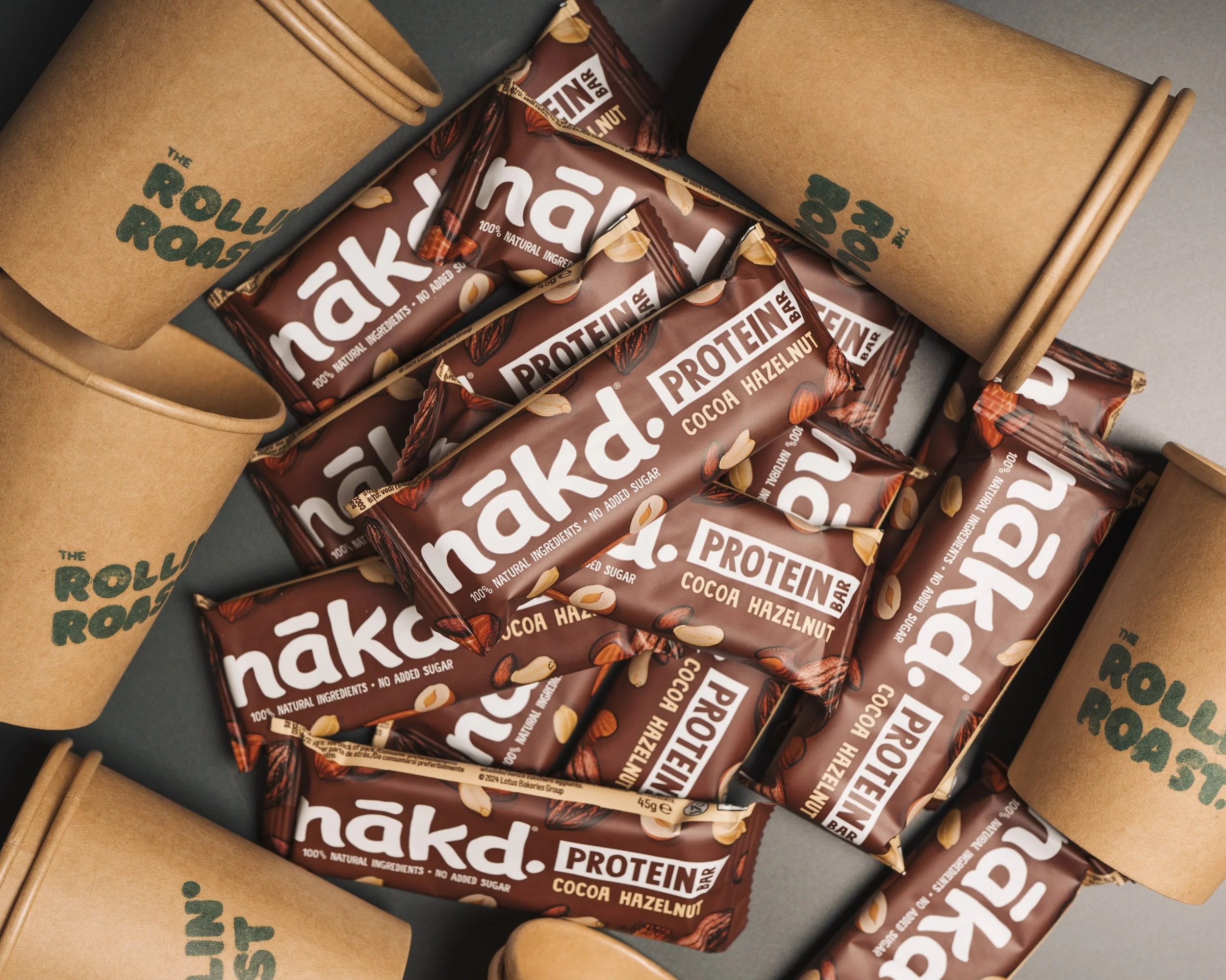 Several chocolate snack bars with cocoa hazelnut flavor are arranged in a box, surrounded by brown paper coffee cups and wooden sticks.