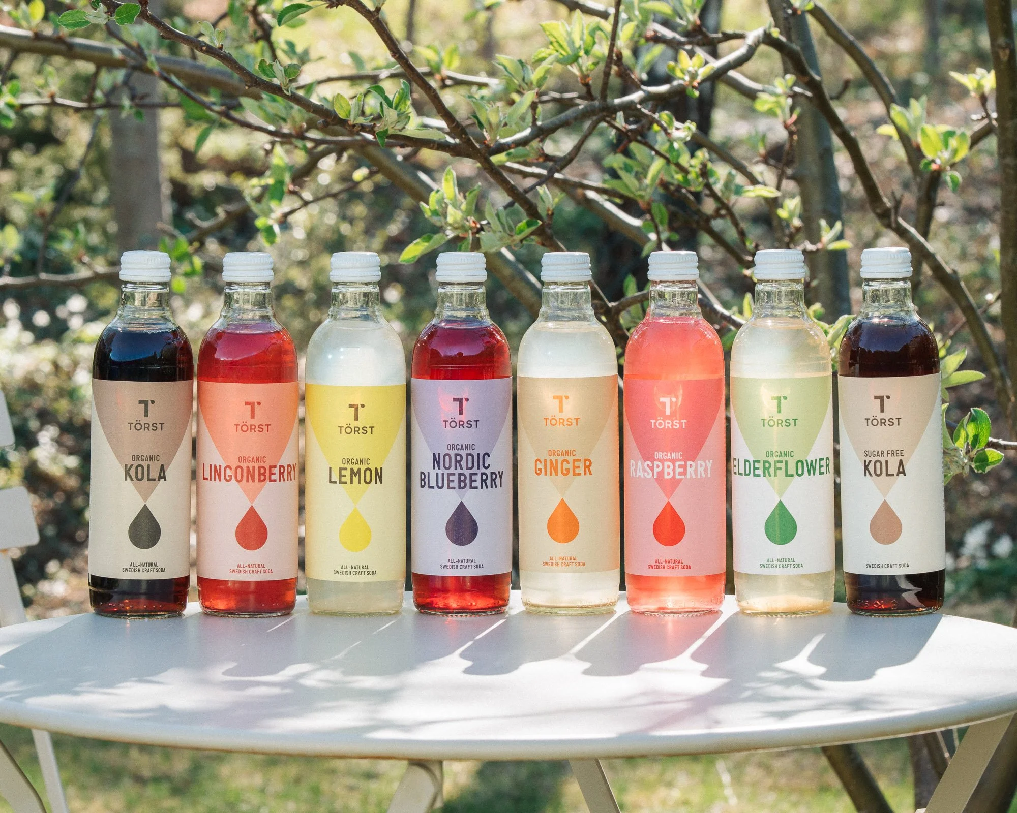Six bottles of oda, each with a different color and flavor, outdoors on a white table against a background of tree branches and leaves. The bottles are labeled flavors including cola, lingonberry, lemon, blueberry, ginger, raspberry, elderflower, and