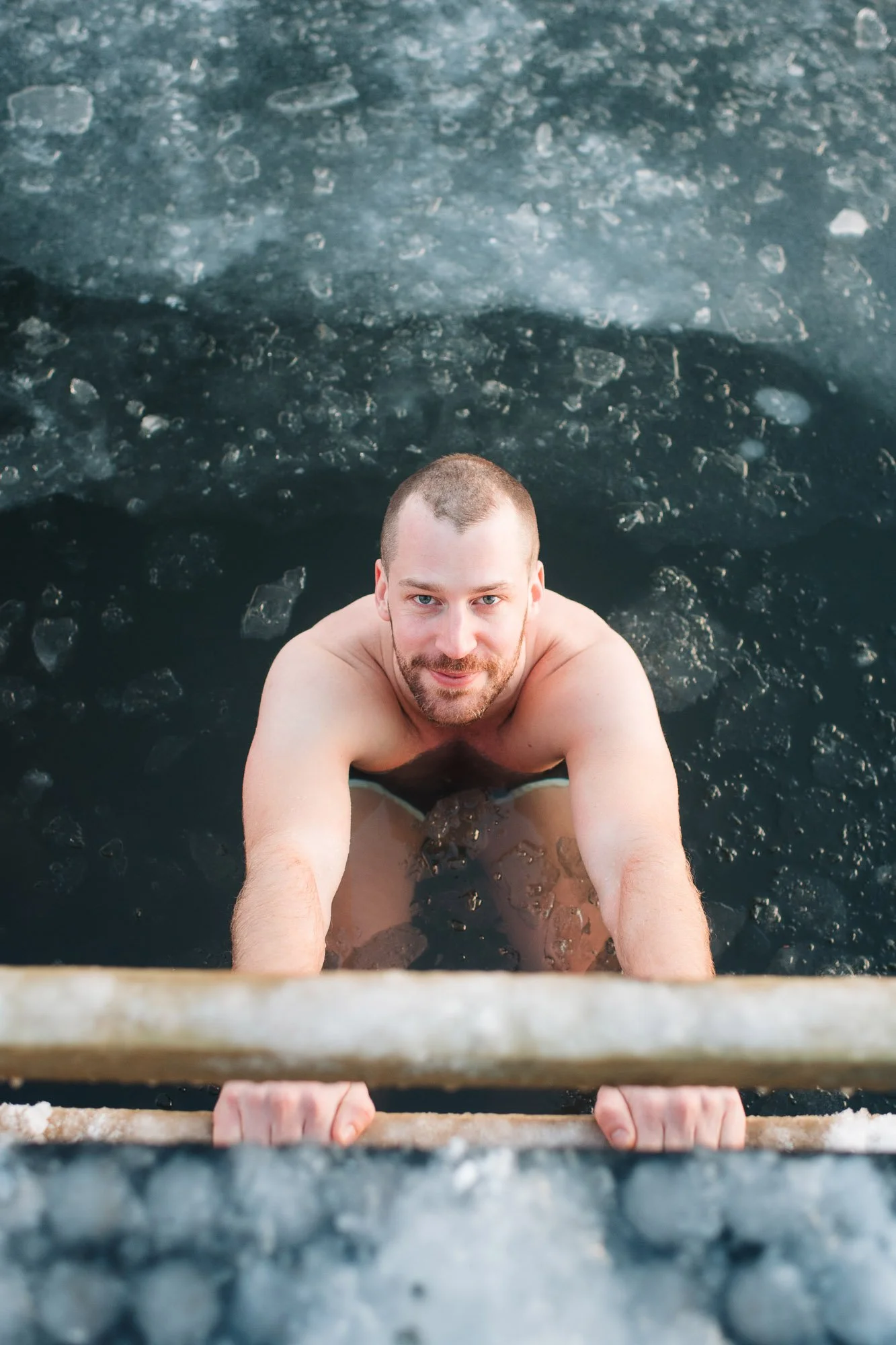 A man with short hair and a beard taking a selfie while climbing out of icy water, holding onto a rusted metal ladder.