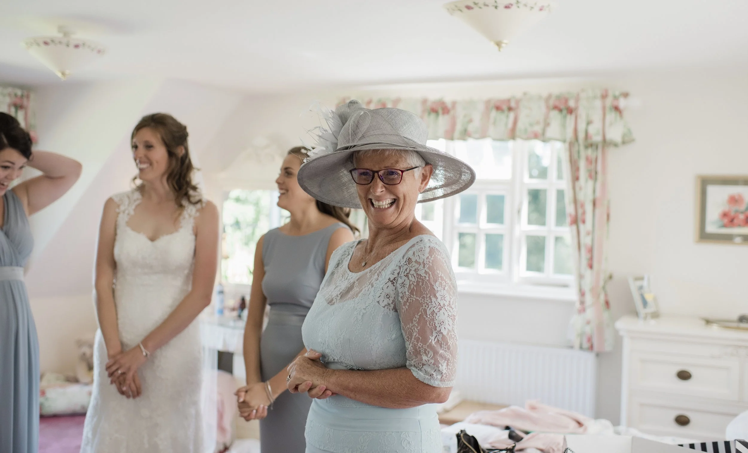 A group of women, including an older woman wearing a large, decorative hat and glasses, smiling and standing in a well-lit room with floral curtains, preparing for a wedding or special event.