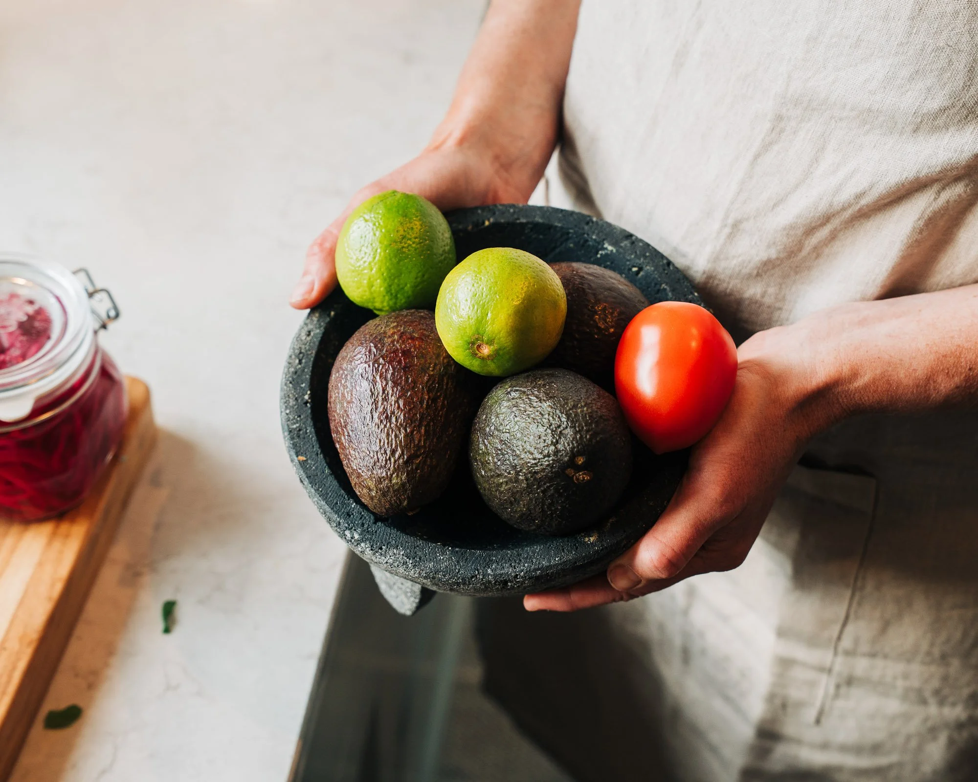 Person holding a round black textured bowl filled with a green lime, a yellow lime, and three avocados.