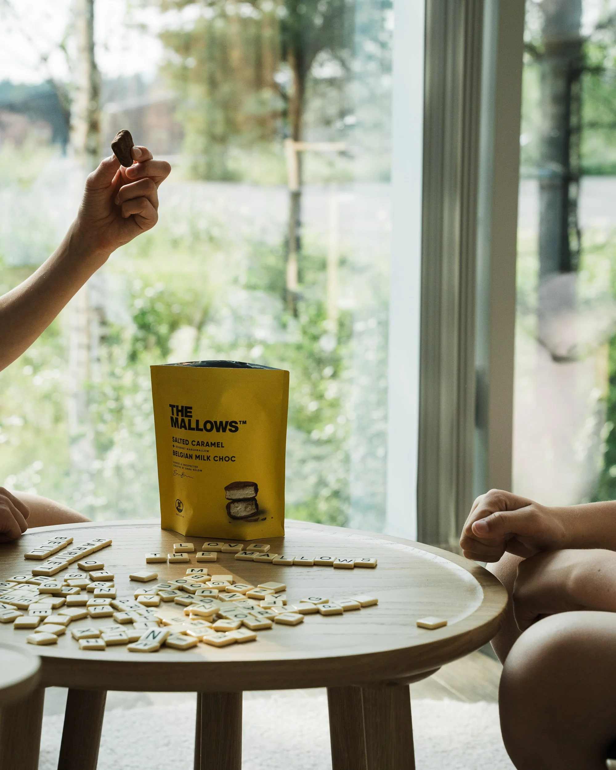 Children playing Scrabble at a table with a bag of The Mallows salt caramel and Belgian milk chocolate candies, near a window showing trees outside.