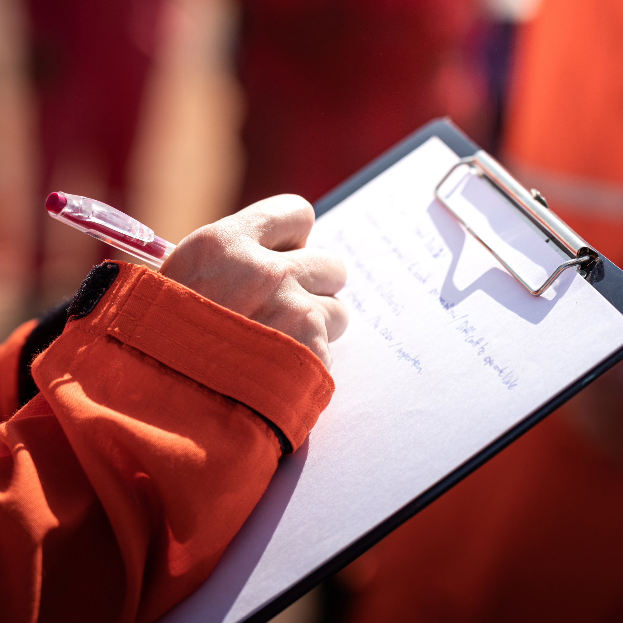 Close-up of a person's hand holding a clipboard with a paper and a pink pen in their hand. The person is wearing an orange glove or sleeve, and the background appears blurred with red and orange tones.