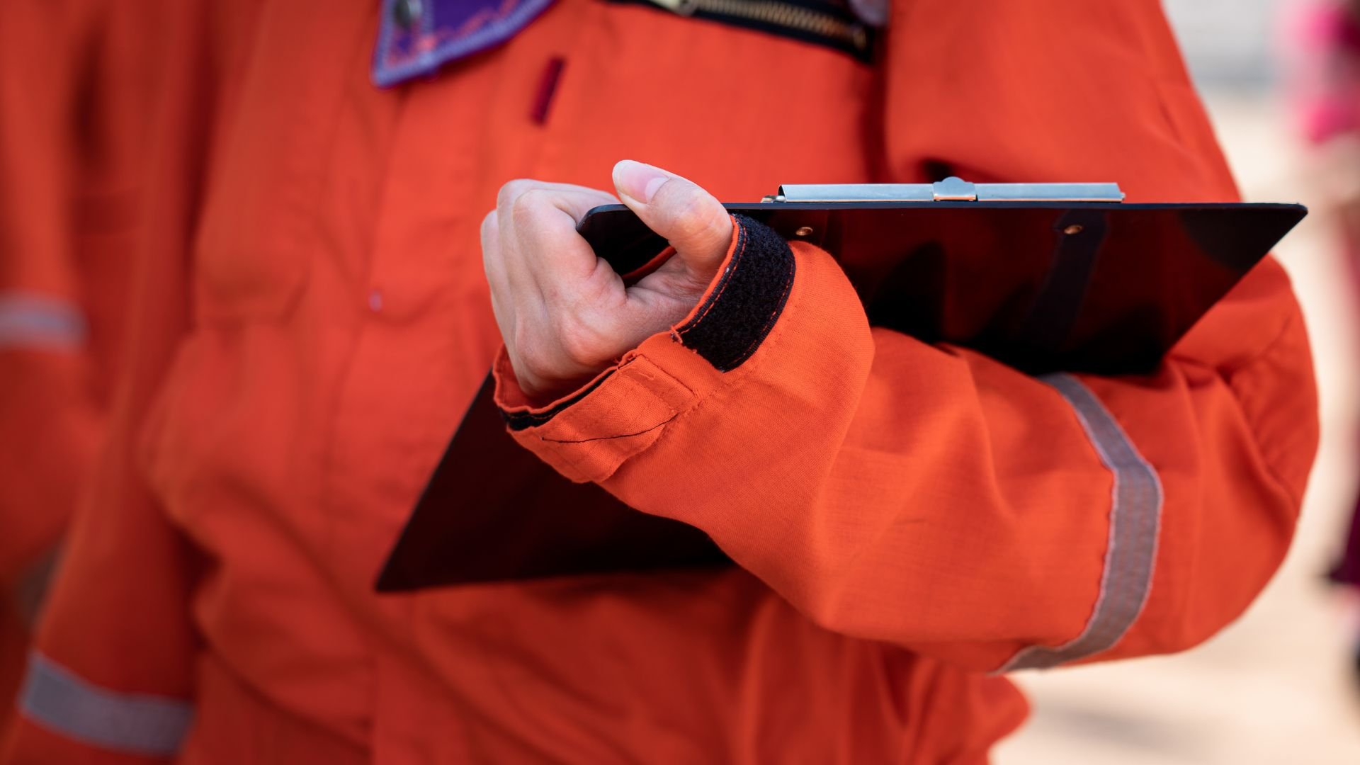 A person wearing an orange safety jacket holding a clipboard, with a thumb over the hand holding the clipboard.