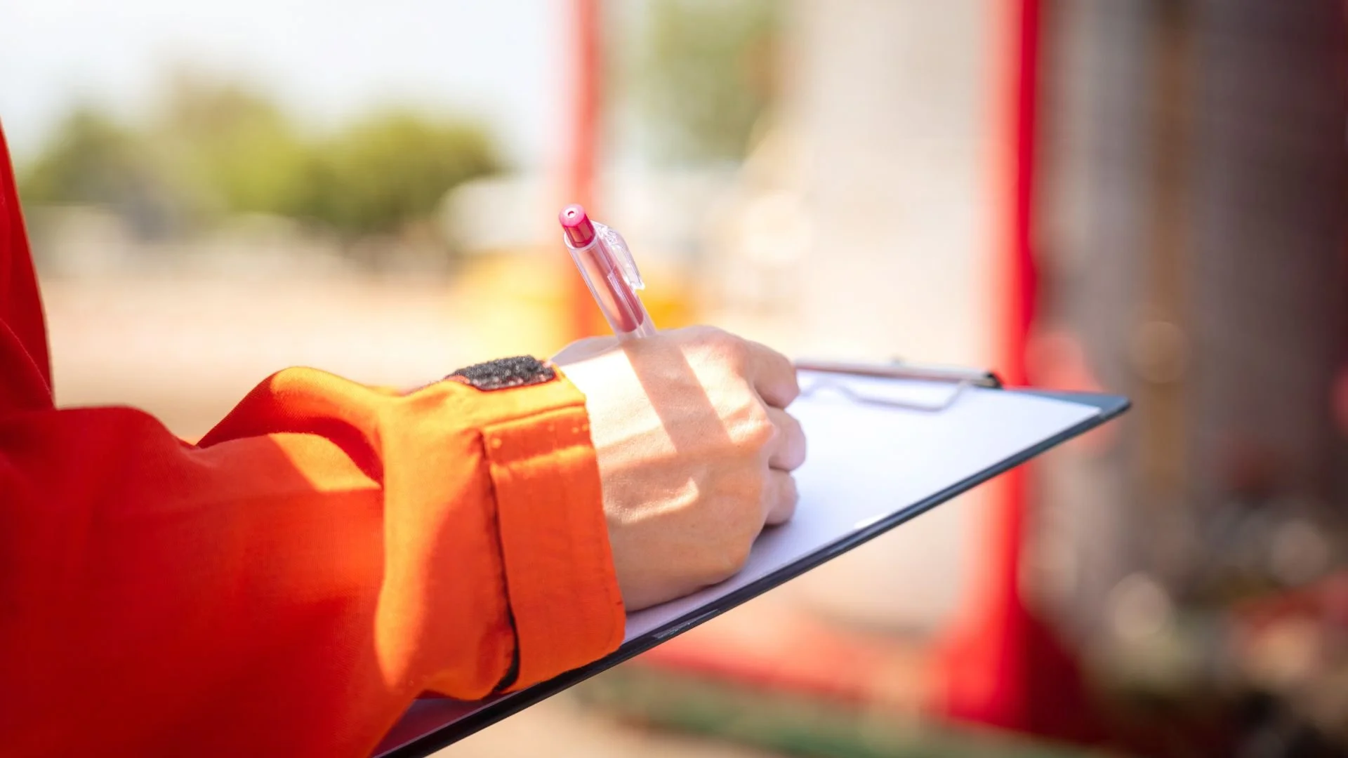 Close-up of a person's hand writing on a clipboard with a pink pen, wearing an orange jacket with a black patch on the sleeve, outdoors with blurred background.