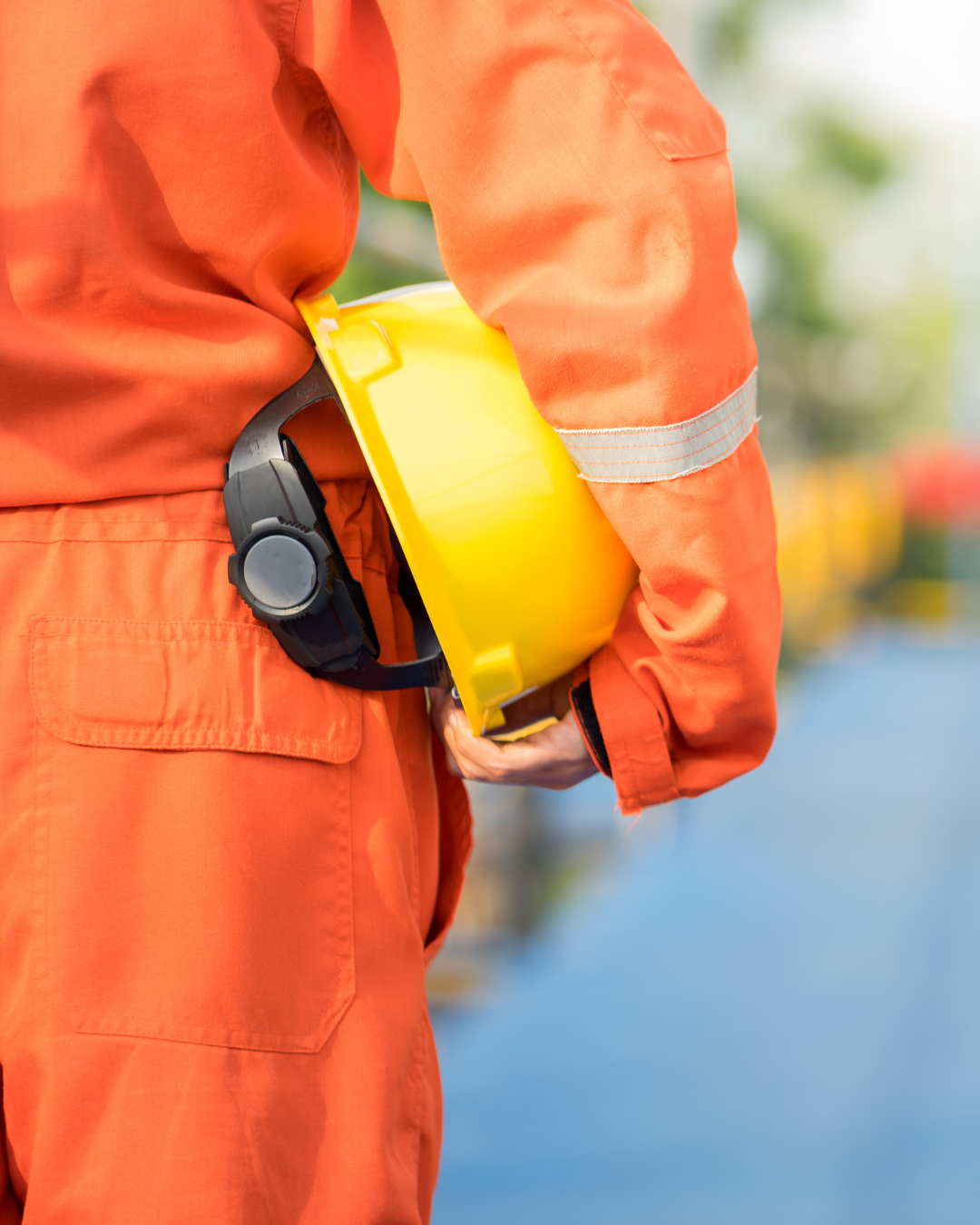 A construction worker wearing orange safety coveralls holding a yellow safety helmet and a hearing protection device.