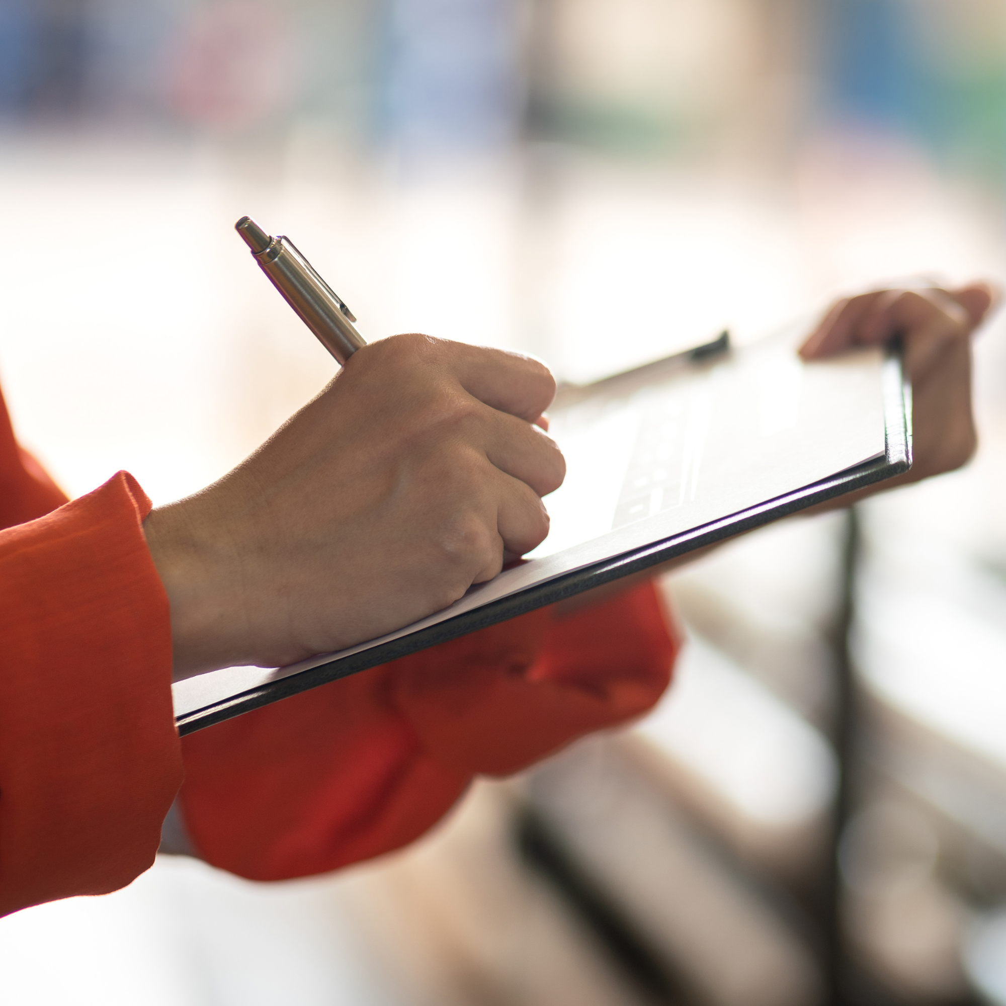 A person holding a metallic pen and writing on a notepad with a colorful blurred background.