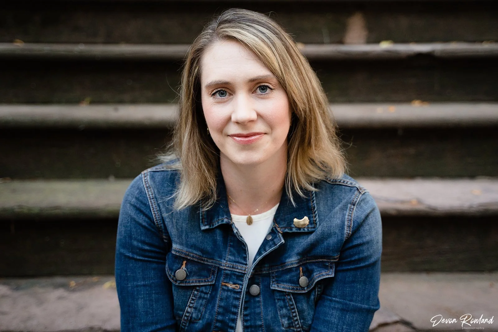 A woman with shoulder-length blonde hair wearing a denim jacket and a white shirt, sitting on outdoor steps, smiling softly.