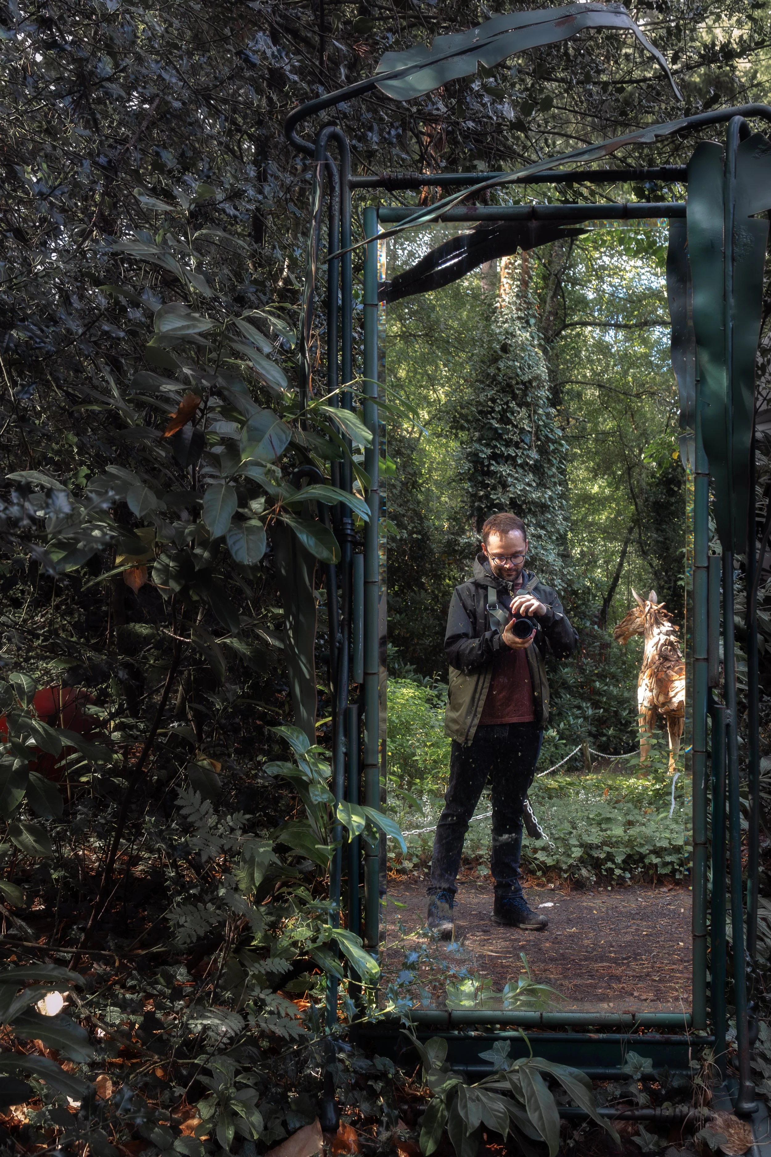 A man with glasses and a backpack standing in a forested area, looking down at his phone, next to a tiger statue or sculpture, with dense green foliage and a glass structure or barrier in the surroundings.