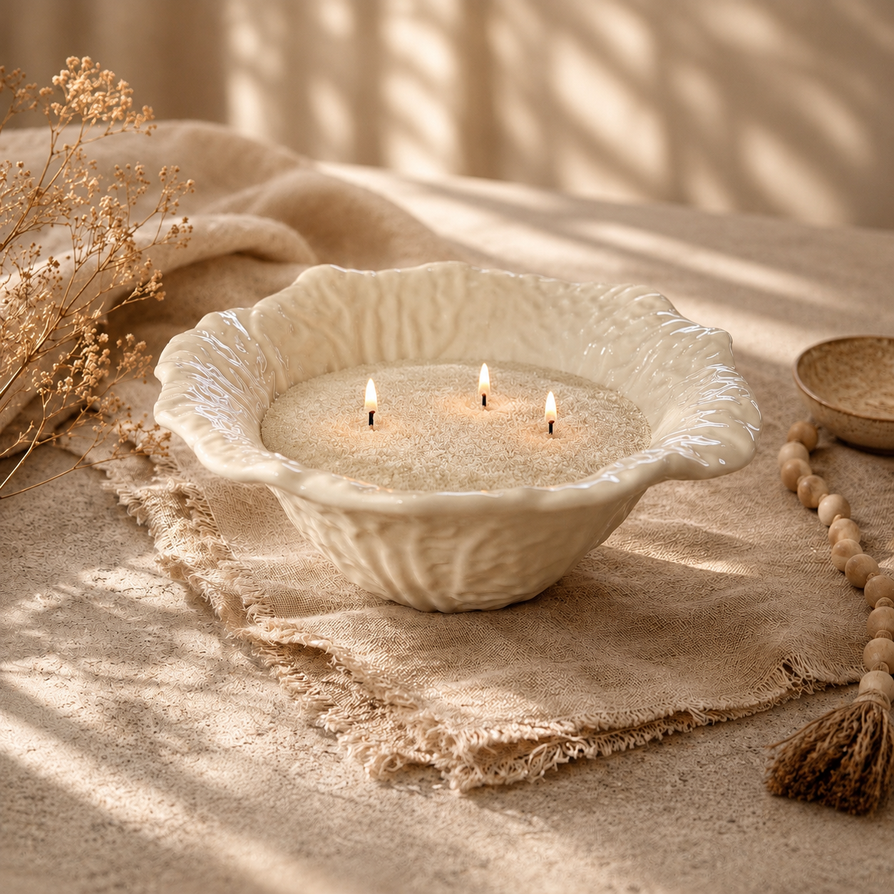 A decorative white ceramic bowl with three burning candles on a textured surface, surrounded by dried flowers, a beaded necklace, and a lace cloth, with soft natural light and shadows creating a warm, cozy atmosphere.