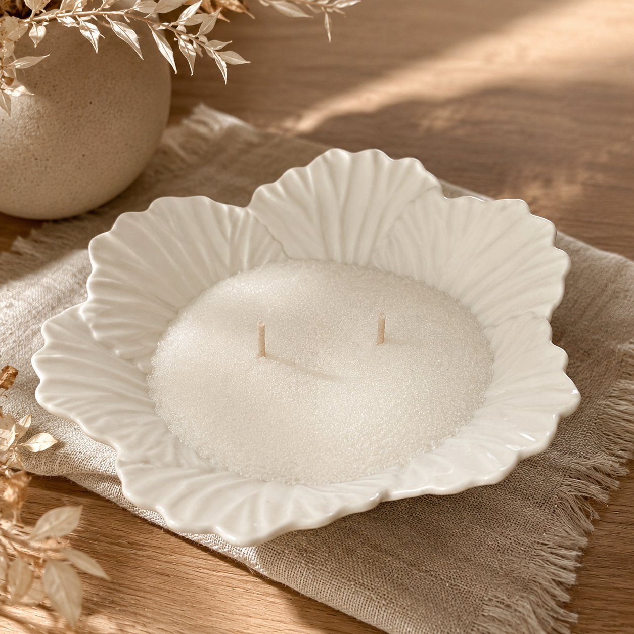 Decorative white candle with two wicks in a scalloped ceramic dish, surrounded by dried plants on a beige cloth on a wooden surface.