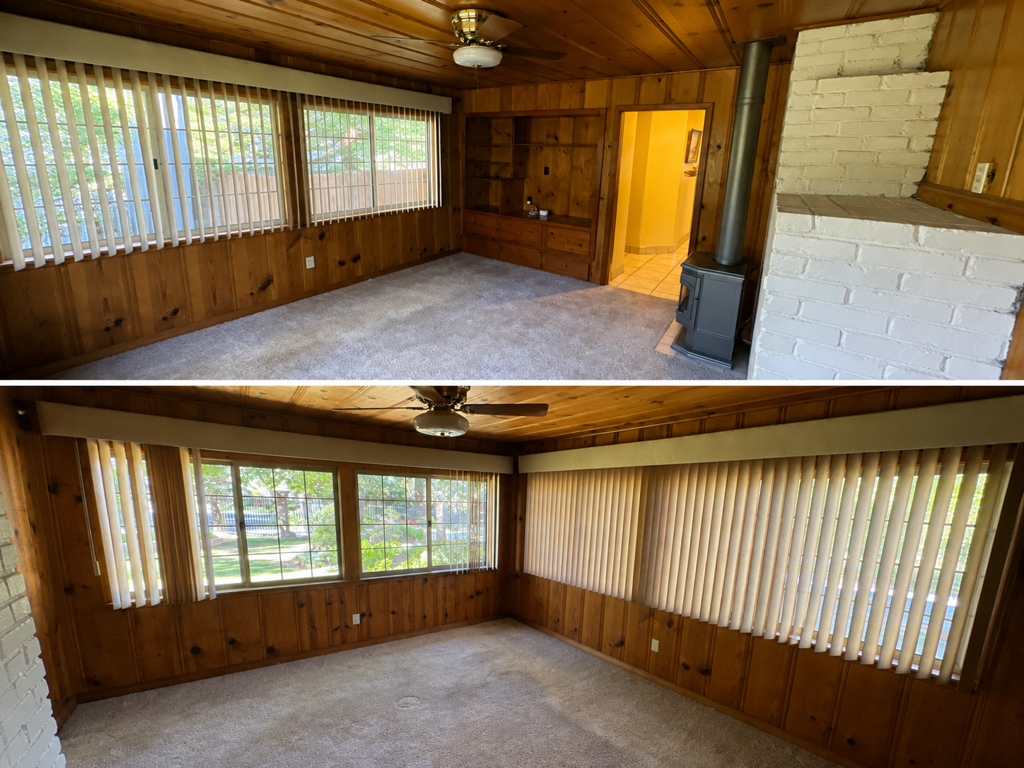 Room with wood-paneled walls, large windows with vertical blinds, carpeted floor, and a wood stove near a white brick wall. The room is empty and has a ceiling fan.