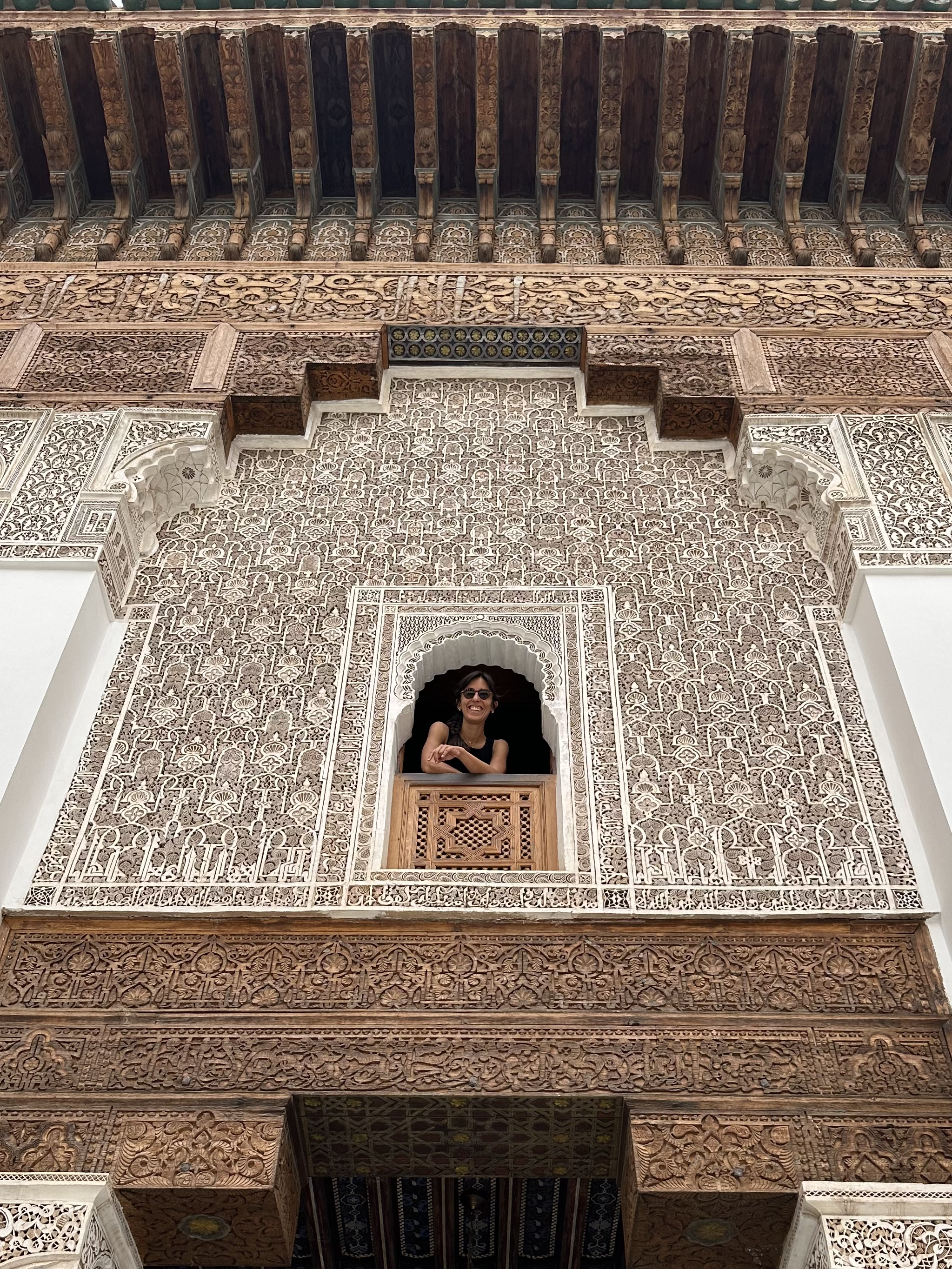 A woman smiling and leaning out of a decorative window in an intricately carved building facade with traditional Moroccan architectural design.